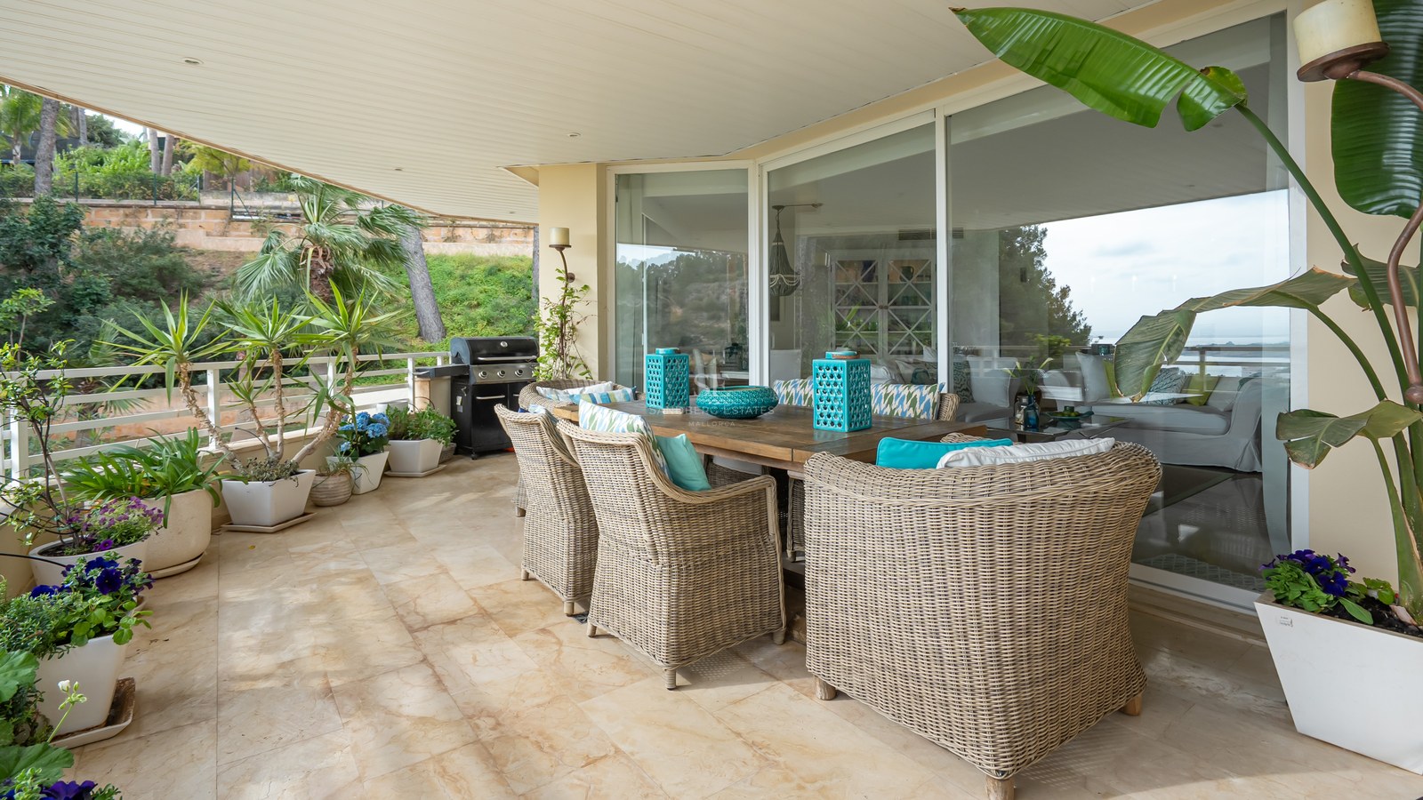 A shaded terrace featuring a wooden dining table, wicker armchairs with turquoise cushions, potted plants, and a grill.