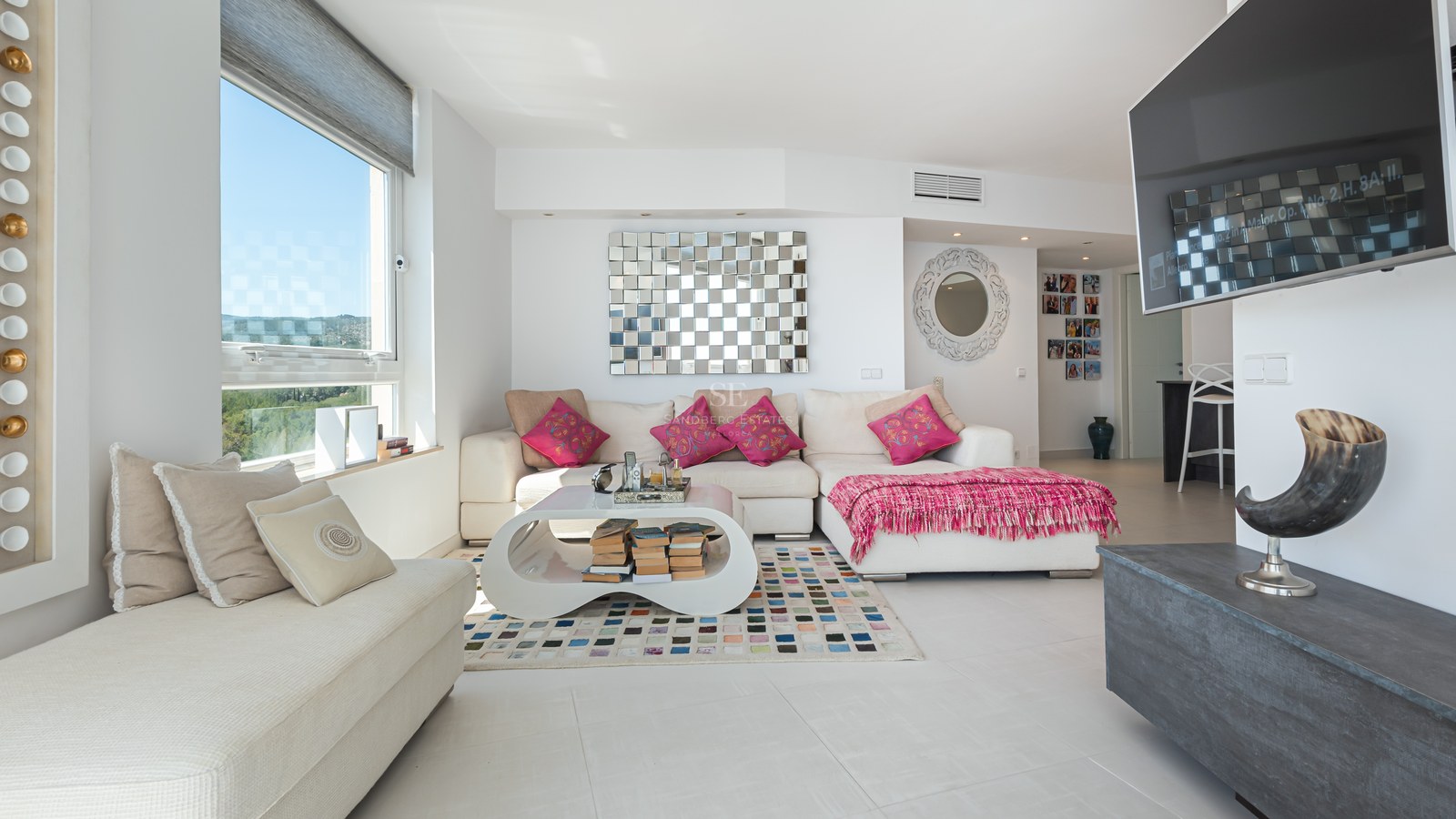 Bright living room with white L-shaped sofa, colorful rug, large window, and wall-mounted television.