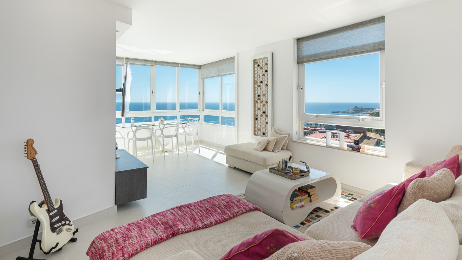 Bright modern living room featuring white furniture, pink accents, and large windows overlooking the Mediterranean sea.