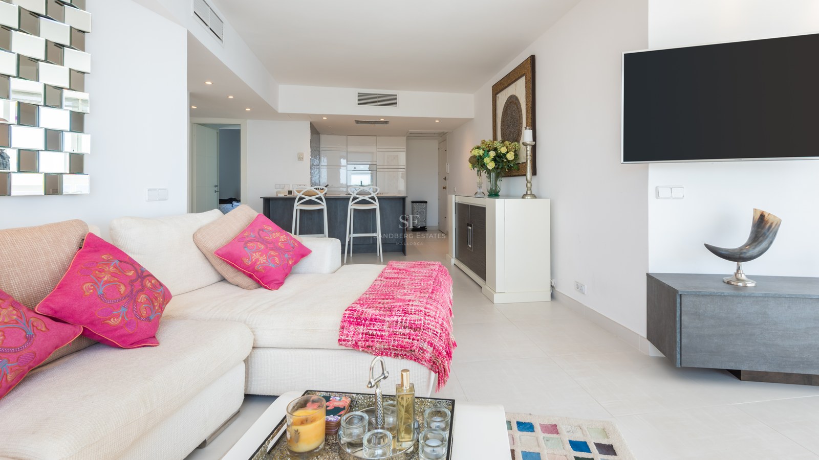 Bright living room with white L-shaped sofa, pink cushions, wall-mounted TV, and an open-plan kitchen in the background.