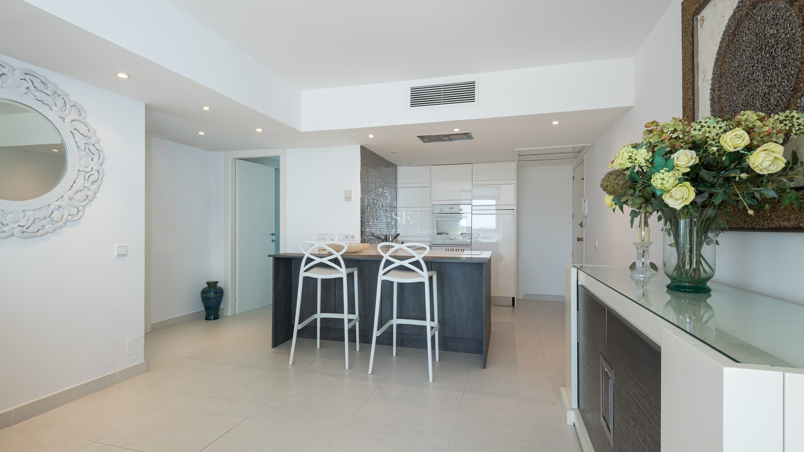 Modern open-plan kitchen featuring a dark grey island, white designer stools, and high-gloss cabinetry.