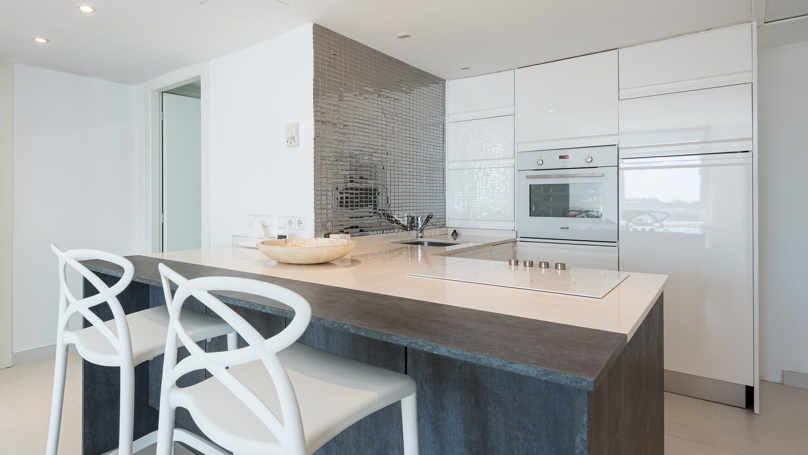 Contemporary white kitchen featuring a dark breakfast bar, white designer chairs, and a silver mosaic backsplash.