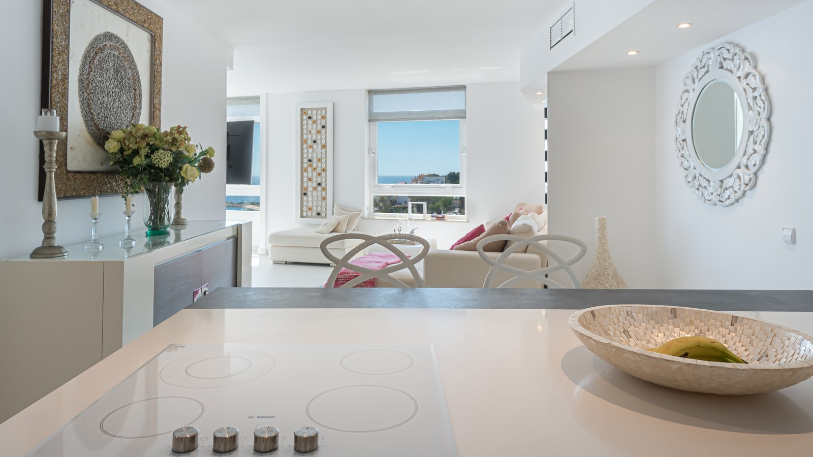 Modern white kitchen island looking into a bright living room with a sea view window and contemporary furniture.
