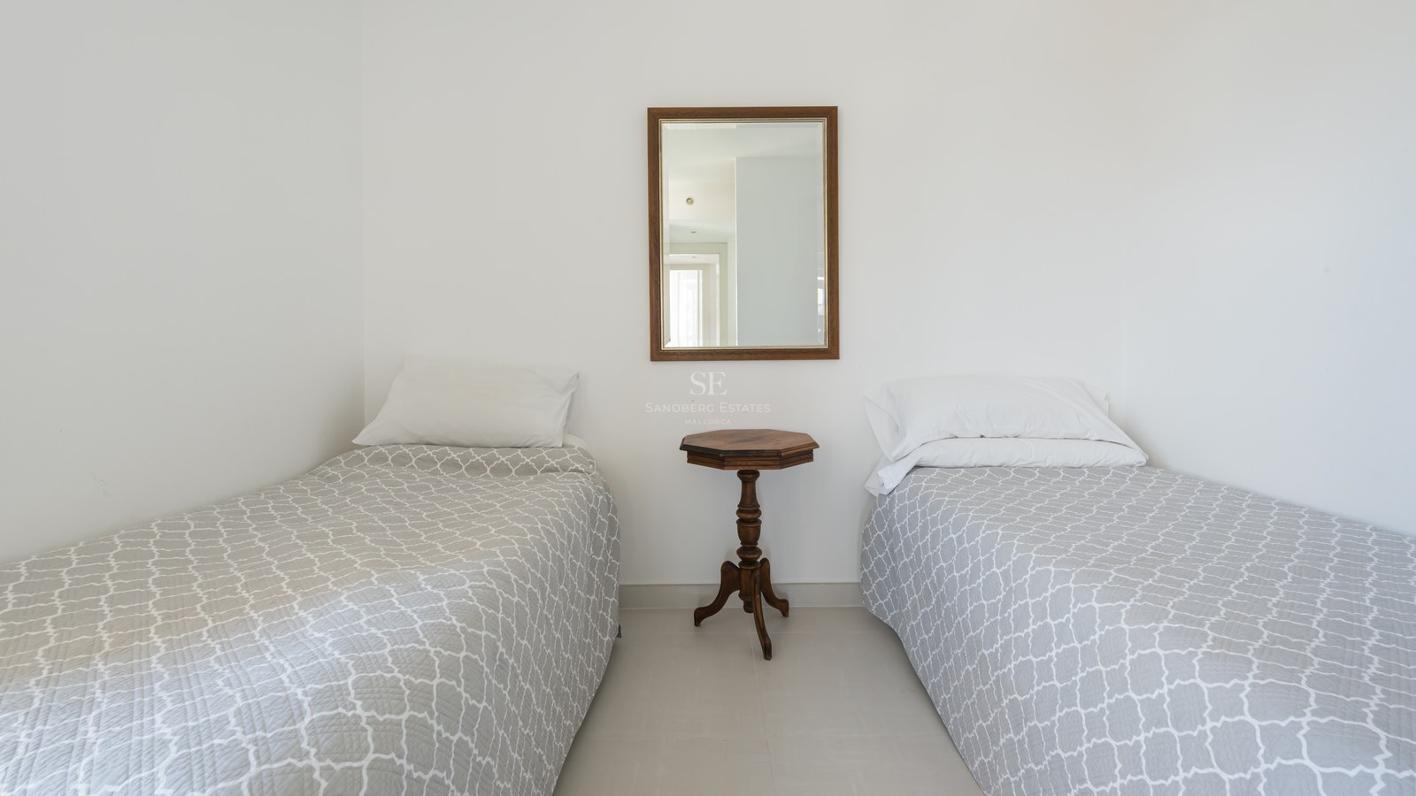White guest bedroom featuring two twin beds with grey patterned covers and a central wooden side table and mirror.
