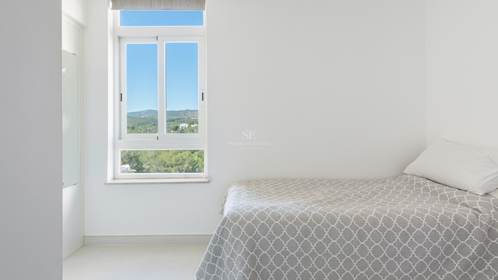 Minimalist white bedroom with a grey patterned bed and a window showing a lush green mountain landscape under a blue sky.