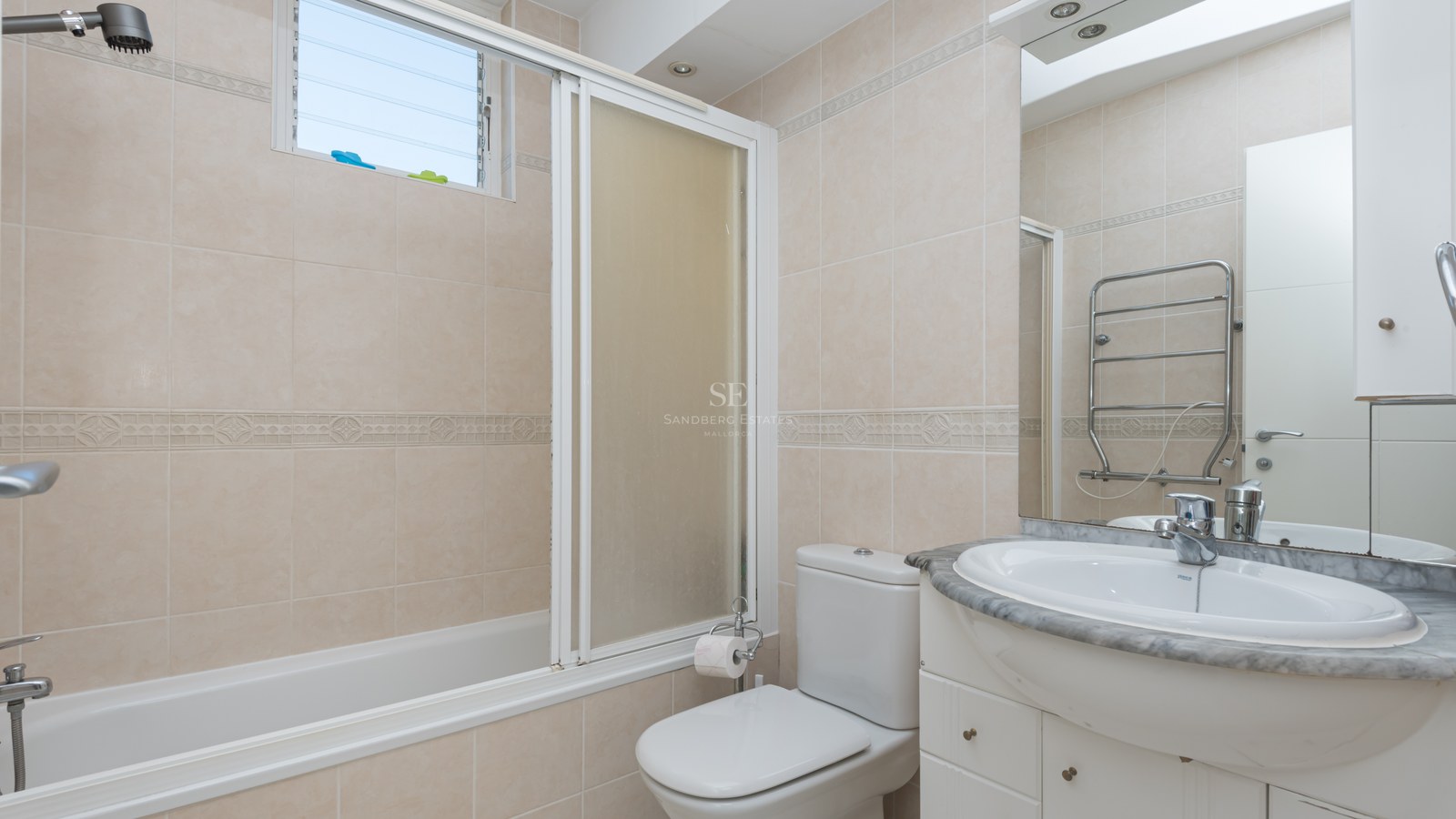 Bright bathroom featuring a bathtub with a screen, a marble vanity unit, a white sink, and neutral ceramic wall tiles.