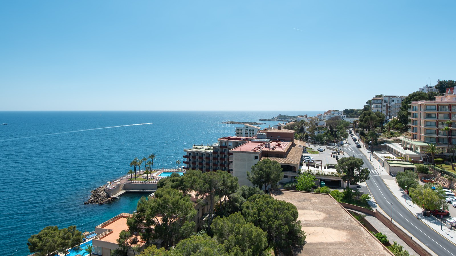 Elevated view of the coastline featuring buildings, pine trees, and the blue sea under a clear sunny sky.
