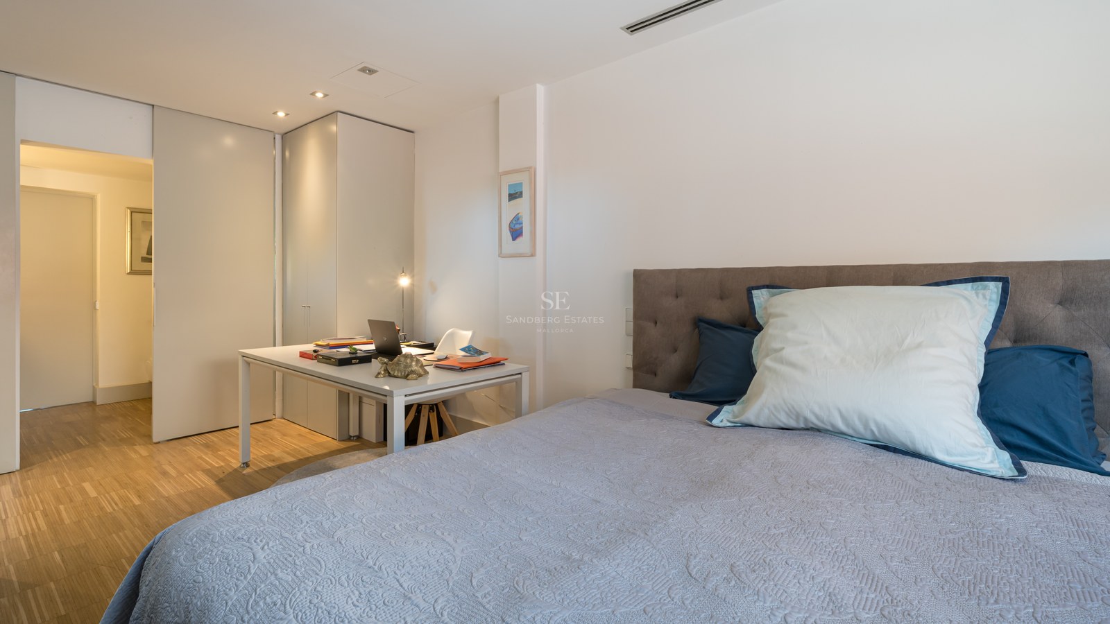 Modernes Schlafzimmer mit grauem Kopfteil, weißen Einbauschränken und Schreibtisch auf Eichenparkett.