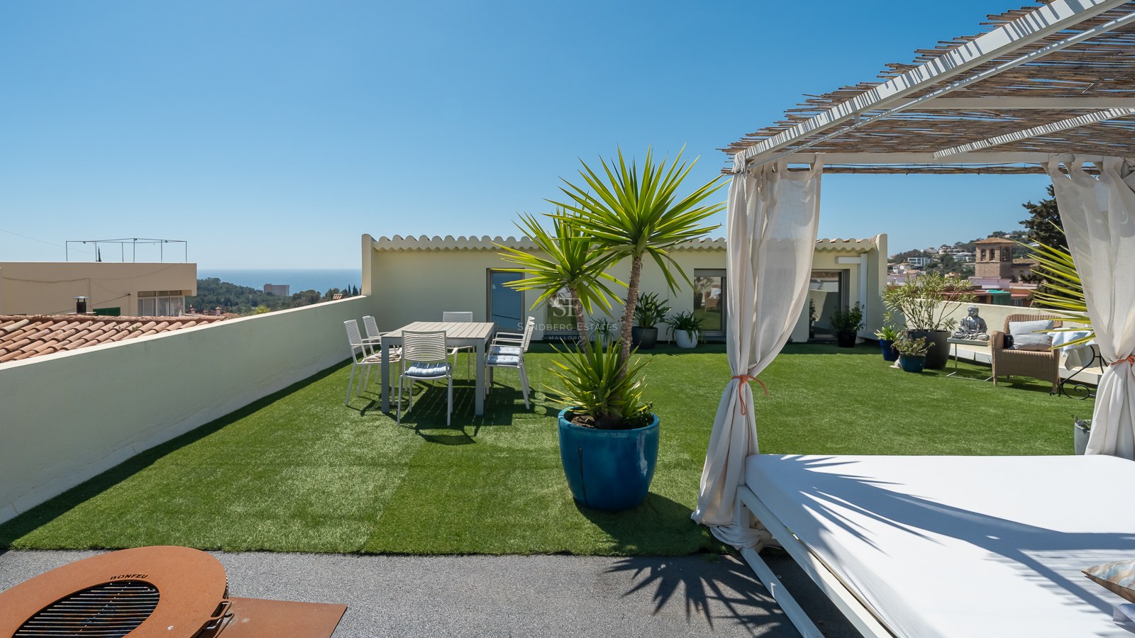 Große Dachterrasse mit Kunstrasen, Esstisch, Daybed unter Bambus-Pergola und Blick auf das Meer.