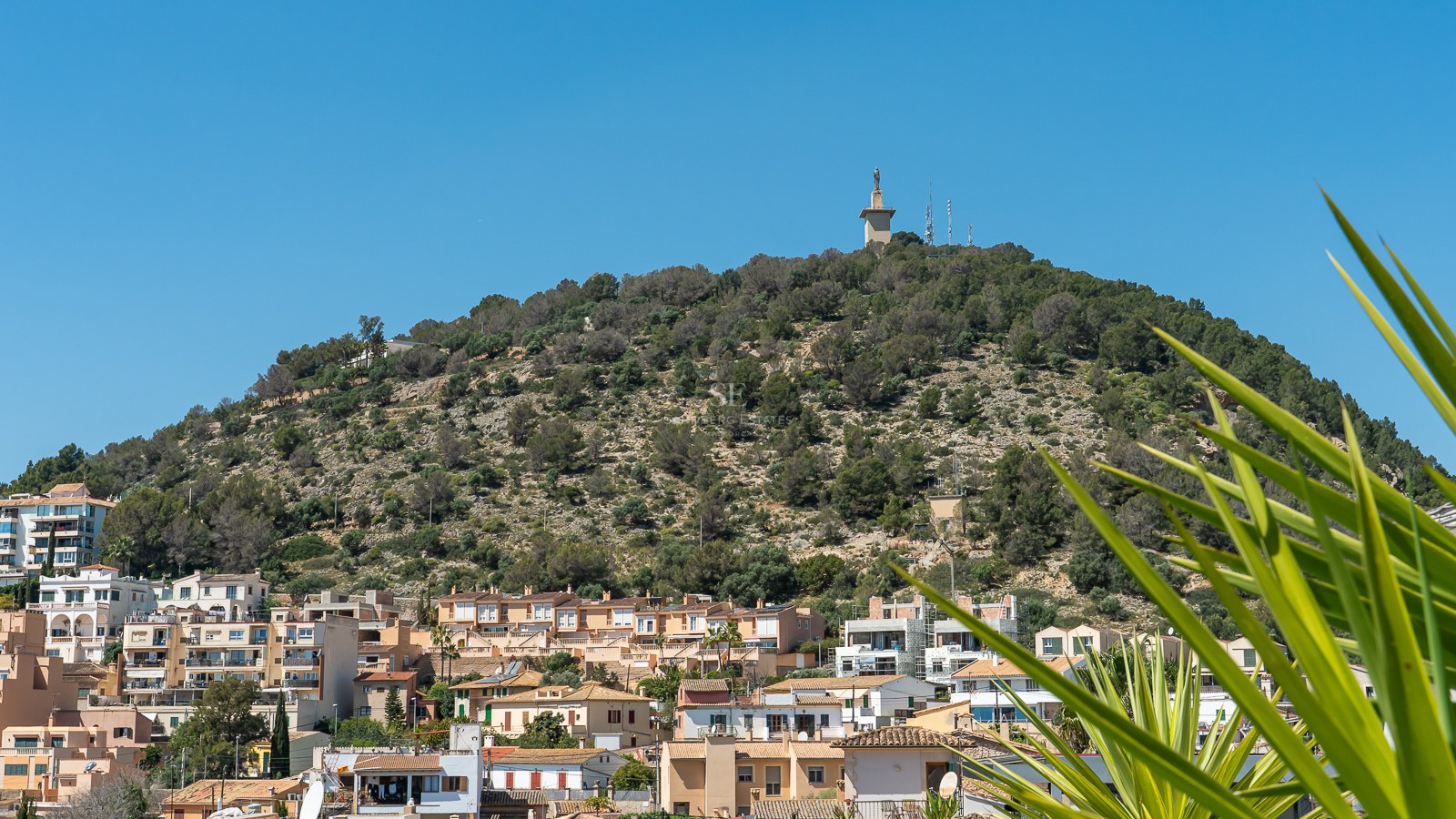 Blick auf einen grünen Hügel mit einem Monument an der Spitze, über einer mediterranen Stadt unter klarem blauem Himmel.
