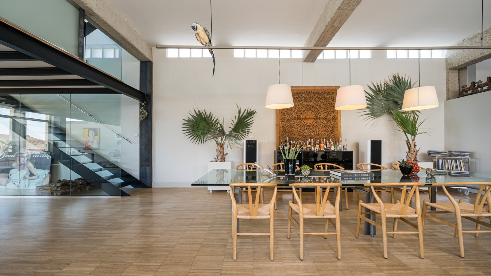 Modernes Esszimmer mit Glastisch, Holzstühlen und Stahltreppe unter hohen Decken mit Sichtbetonbalken.
