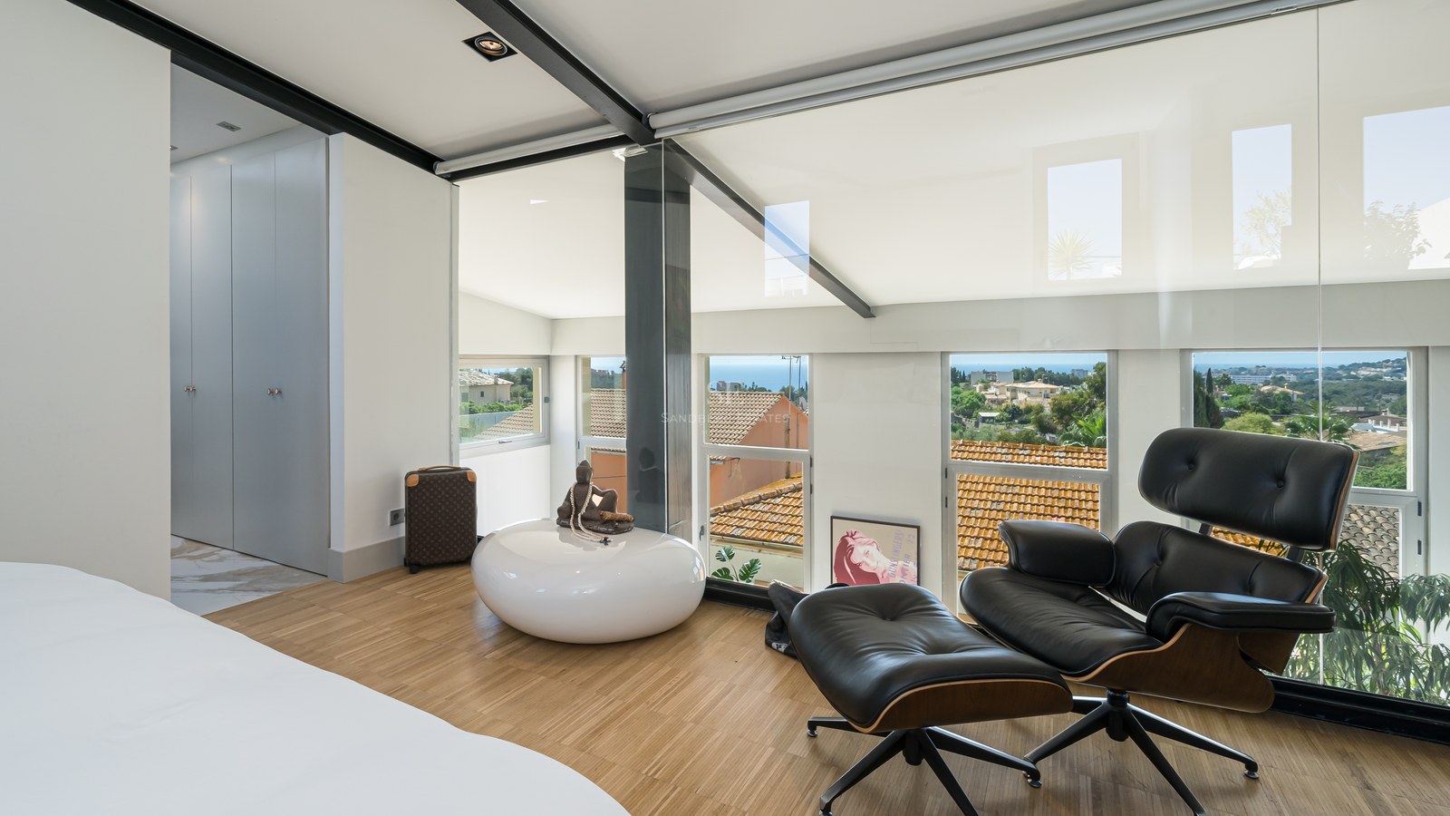 Modernes Schlafzimmer mit Eames Lounge Chair, Eichenparkett und Glasfronten mit Blick auf das Meer und die Stadt.