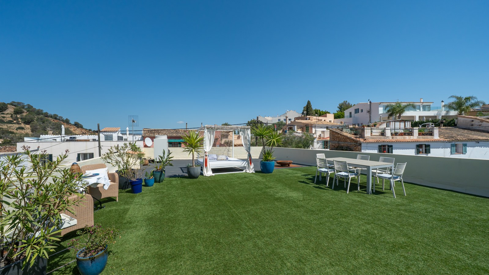 Dachterrasse mit Kunstrasen, weißem Balinesischem Bett, Essbereich im Freien und Blick auf weiße Dorfhäuser.