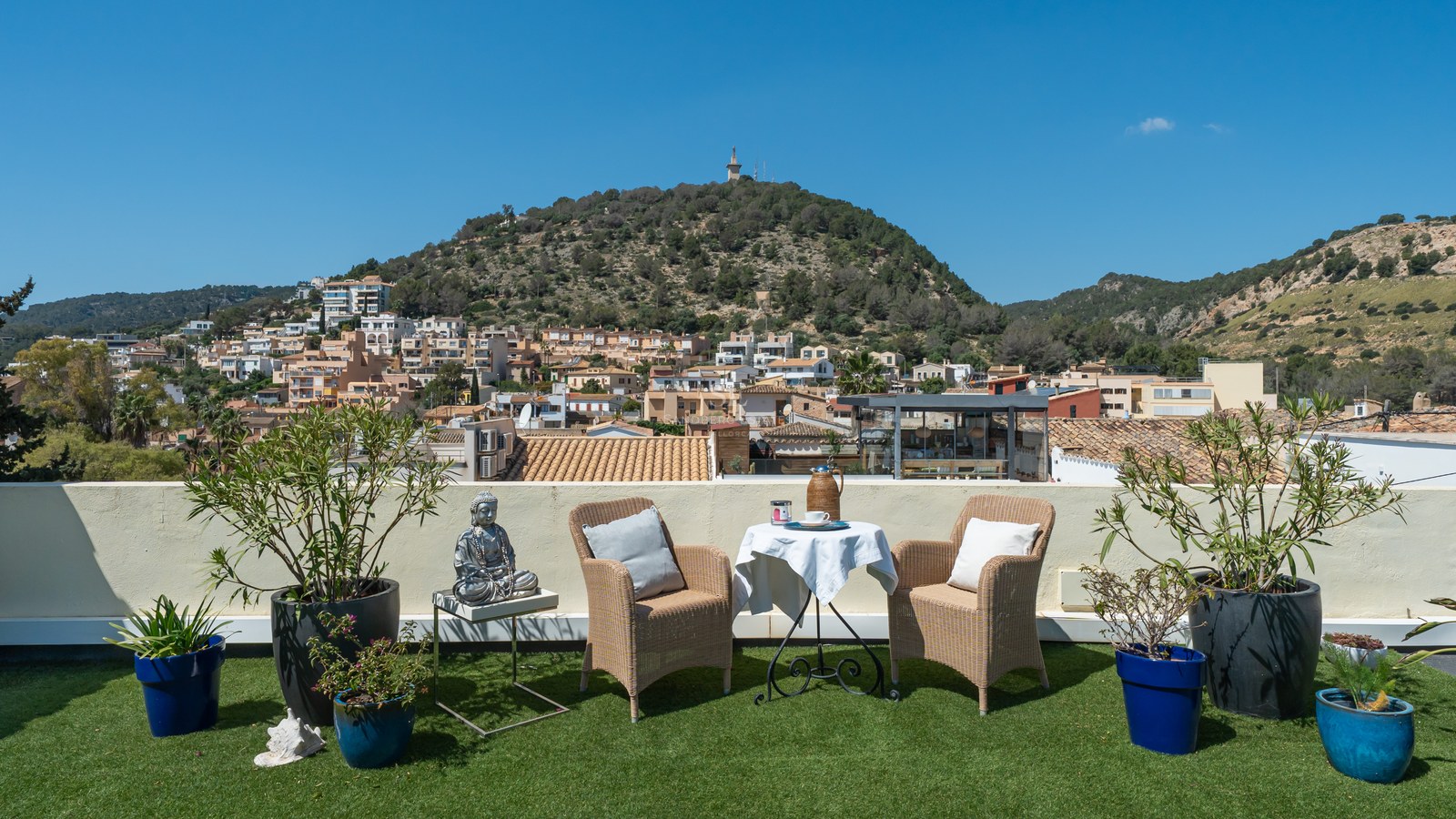 Dachterrasse mit Korbsesseln, Beistelltisch und Pflanzen mit Blick auf ein mediterranes Dorf und einen bewaldeten Hügel.