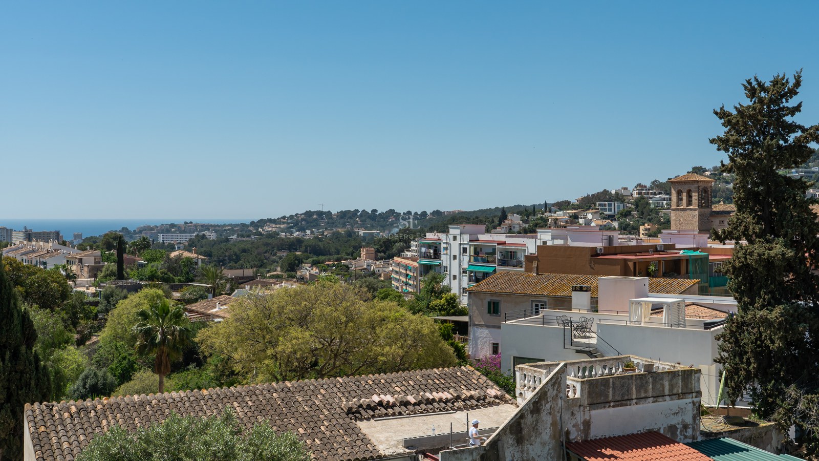 Blick über mediterrane Dächer, grüne Hügel und das ferne blaue Meer unter klarem Himmel.