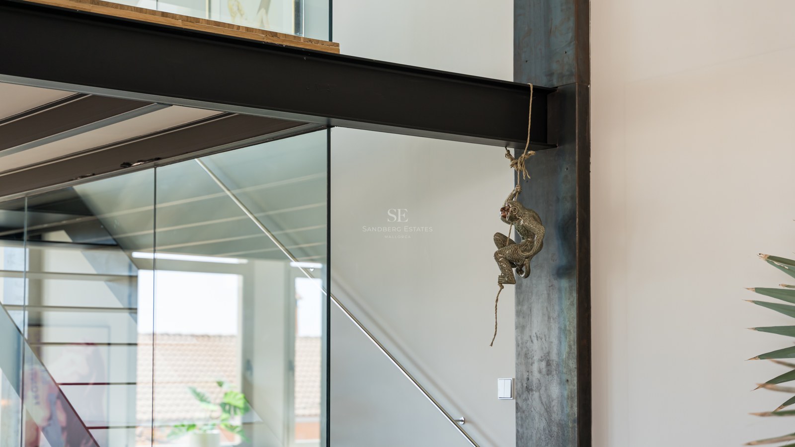 Schwarzer Stahlträger mit dekorativer Affenskulptur an einem Seil neben Glasgeländern.