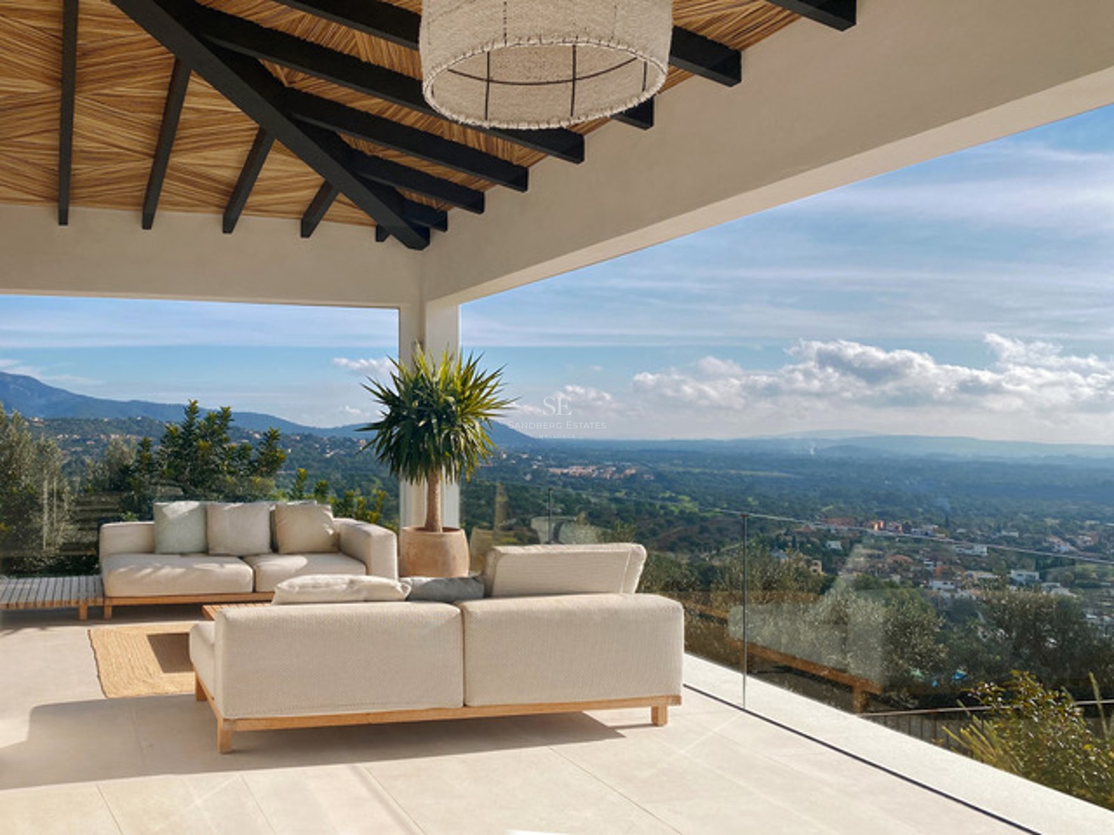 Moderne Terrasse mit cremefarbenen Sofas, Holzbalkendecke und Glasgeländer mit weitem Blick über das Tal.