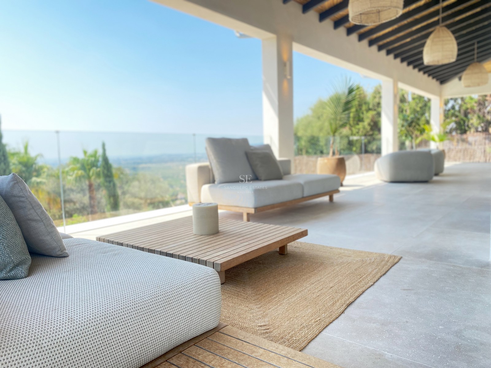 Moderne überdachte Terrasse mit grauen Loungemöbeln, Teakholz und Glasgeländern mit Blick auf ein grünes Tal.