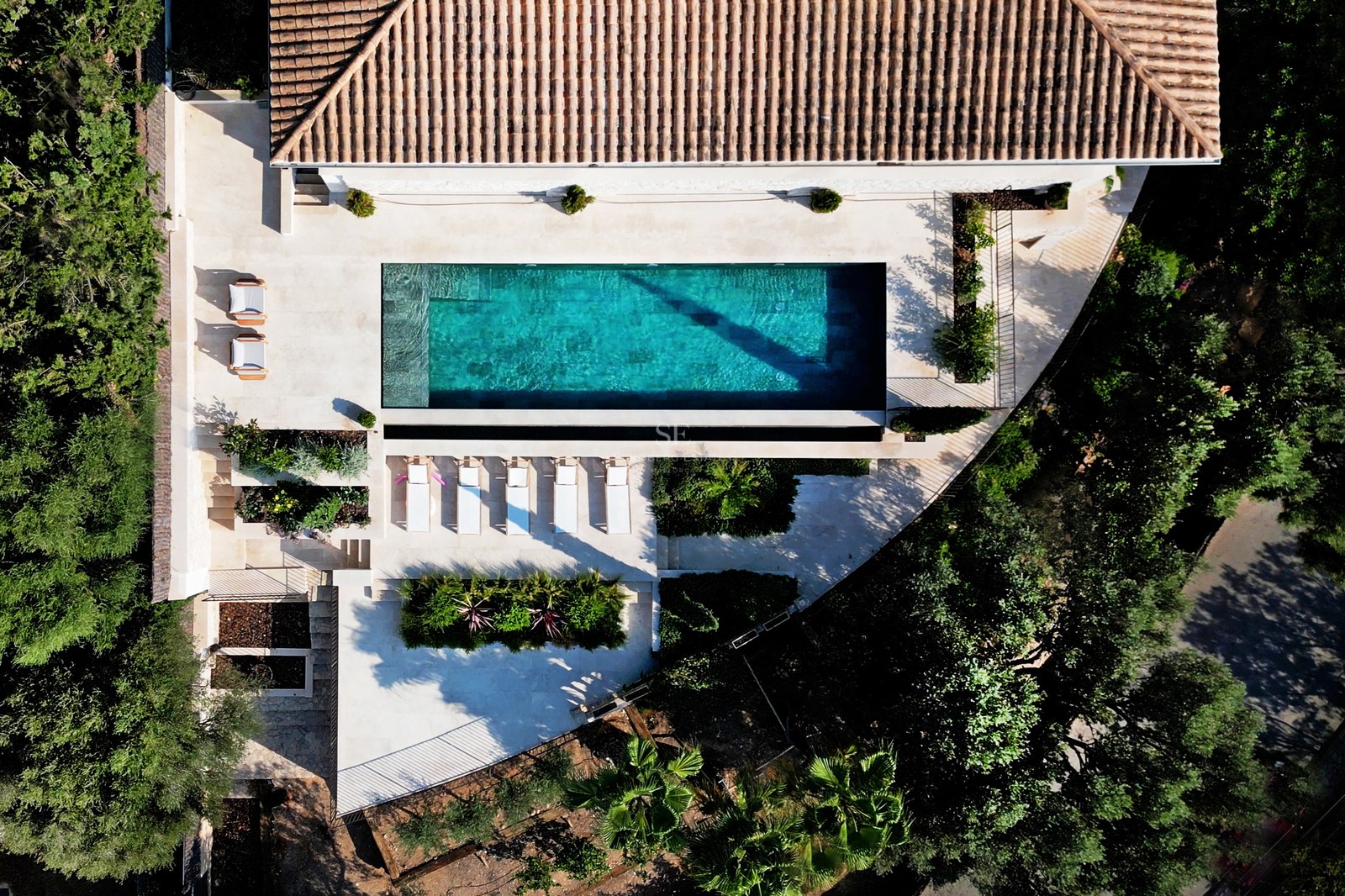 Draufsicht auf einen türkisfarbenen Pool, umgeben von Steinterrassen, Sonnenliegen und üppiger mediterraner Vegetation.