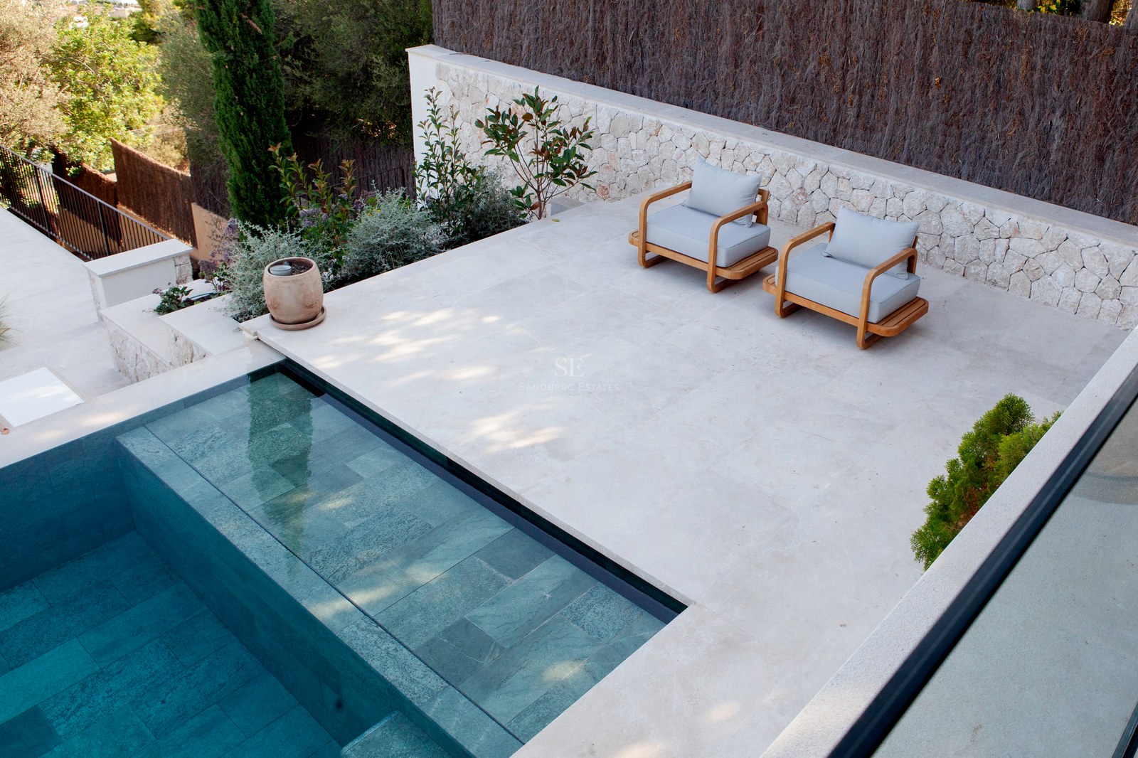 Erhöhte Ansicht eines türkis gefliesten Pools neben einer hellen Steinterrasse mit zwei Designer-Holzsitzsesseln.