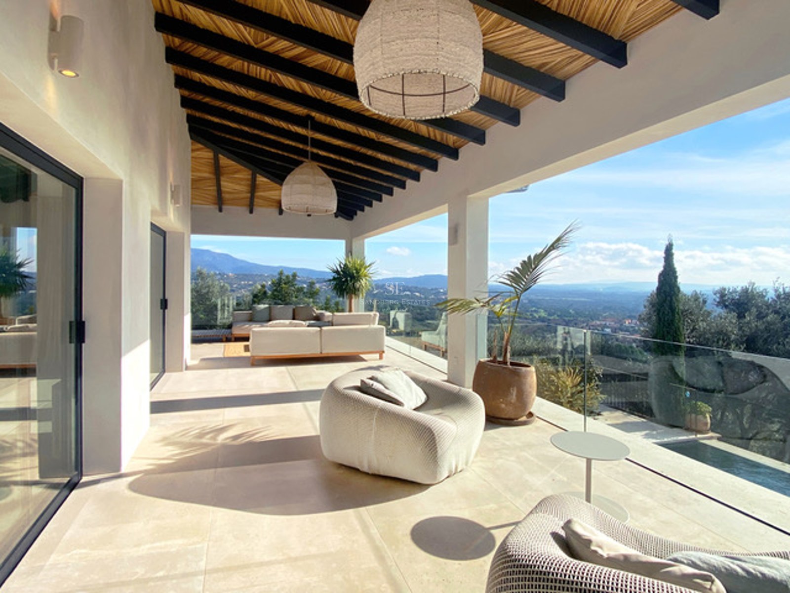 Überdachte moderne Terrasse mit beigen Loungemöbeln, Glasgeländern und weitem Blick auf ein grünes Tal und Berge.