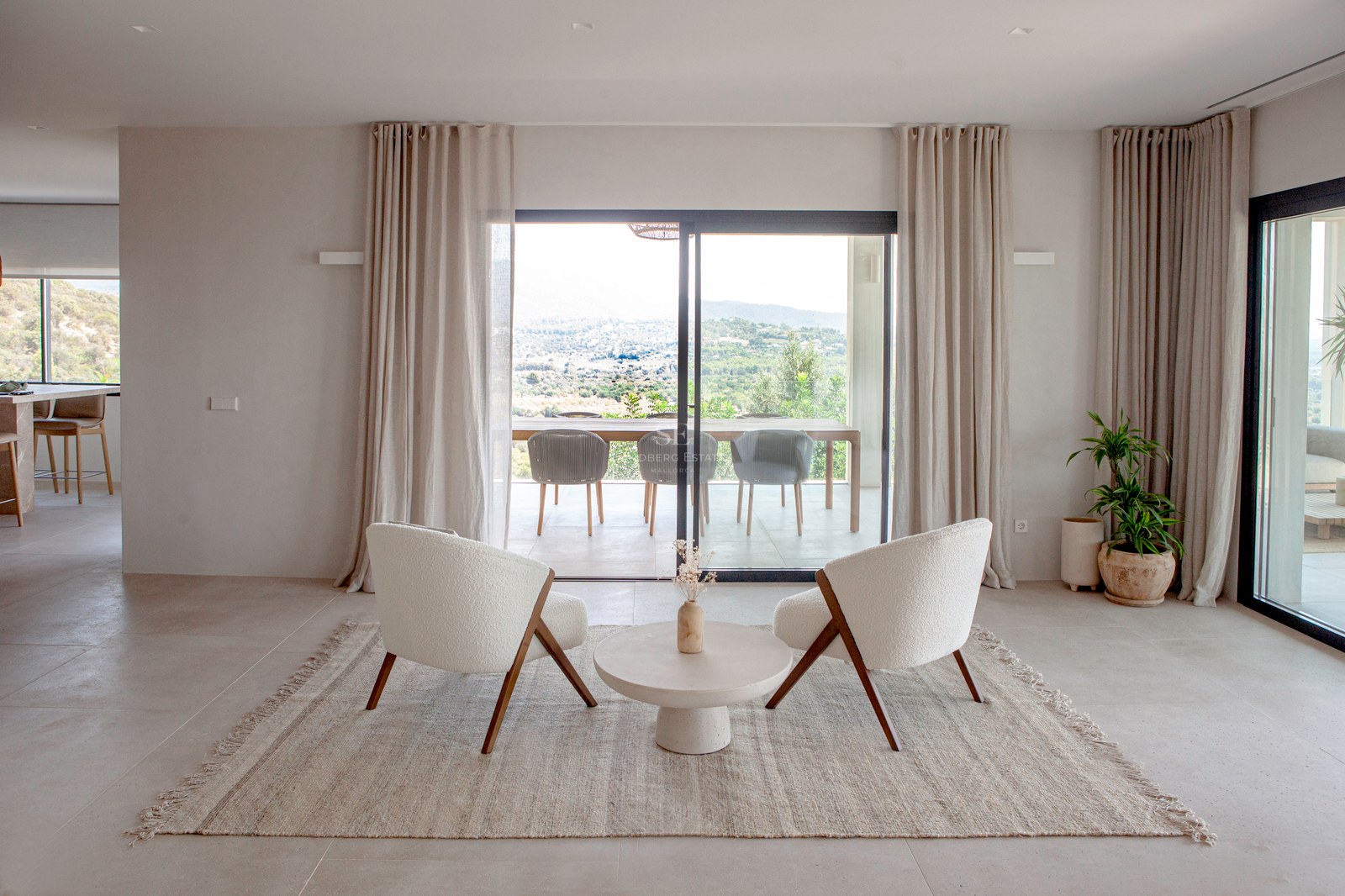 Modernes Wohnzimmer mit zwei cremefarbenen Sesseln, Juteteppich und bodentiefen Fenstern mit Blick auf die Berge.