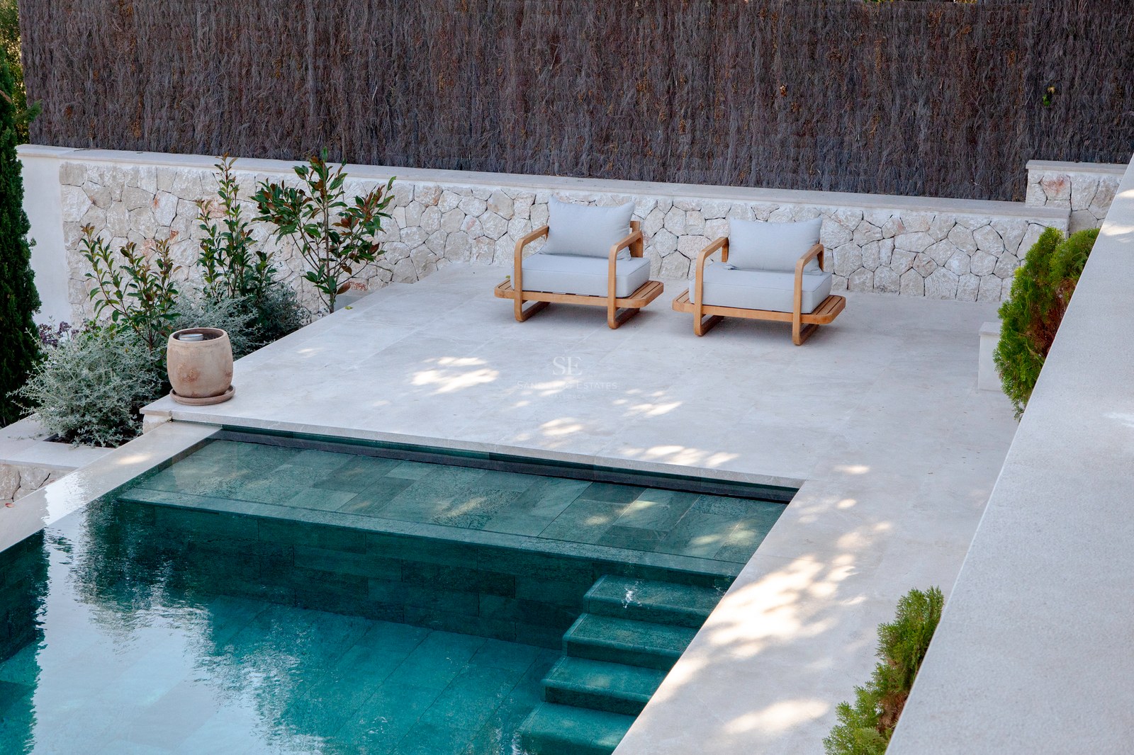 Draufsicht auf einen modernen Pool mit türkisfarbenen Fliesen neben einer Steinterrasse mit zwei Holzsesseln.