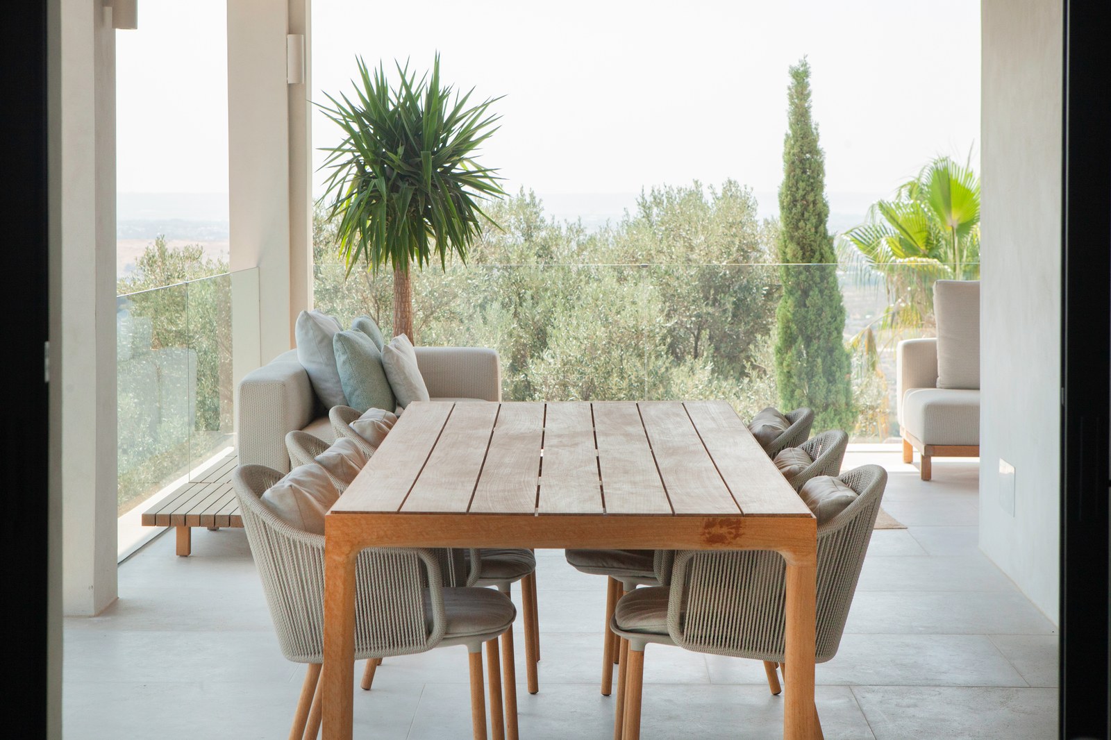 Moderne überdachte Terrasse mit großem Holztisch, Korbstühlen und Blick auf mediterrane Vegetation.