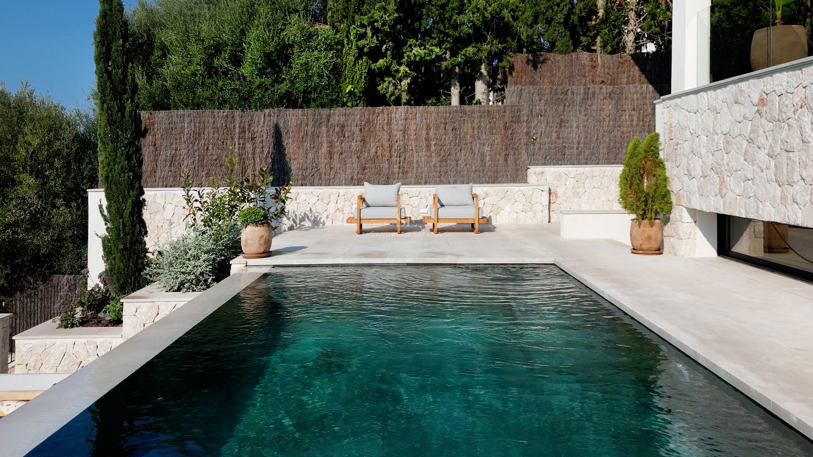 Türkisfarbener Pool neben weißen Natursteinmauern mit Holzliegen und mediterranen Pflanzen im Hintergrund.