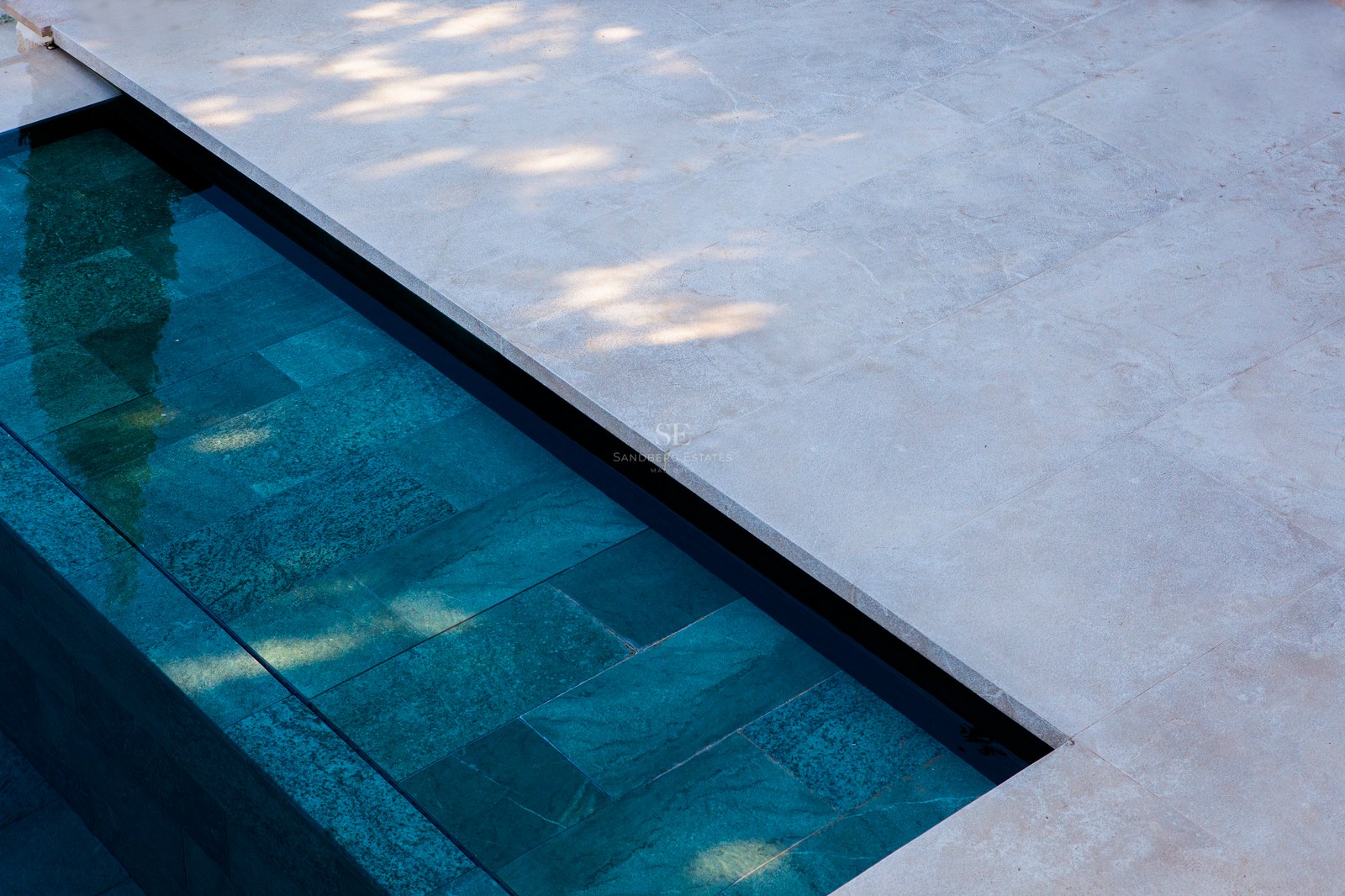 Nahaufnahme eines modernen Pools mit türkisblauen Steinfliesen und hellem, sandfarbenem Naturstein-Terrassenbelag.