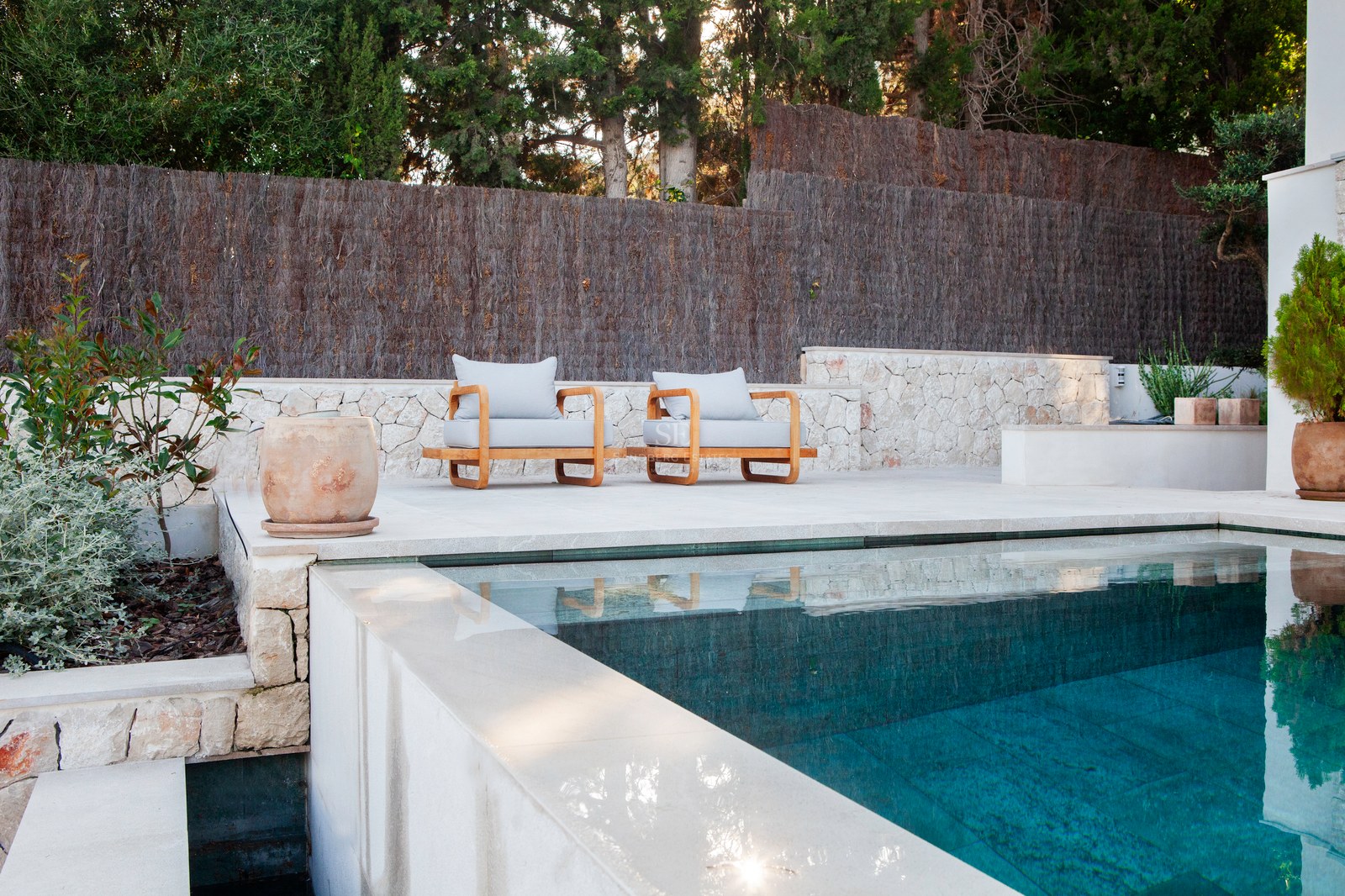 Infinity-Pool mit türkisblauem Wasser neben einer Kalksteinterrasse mit zwei Designer-Holzsesseln.