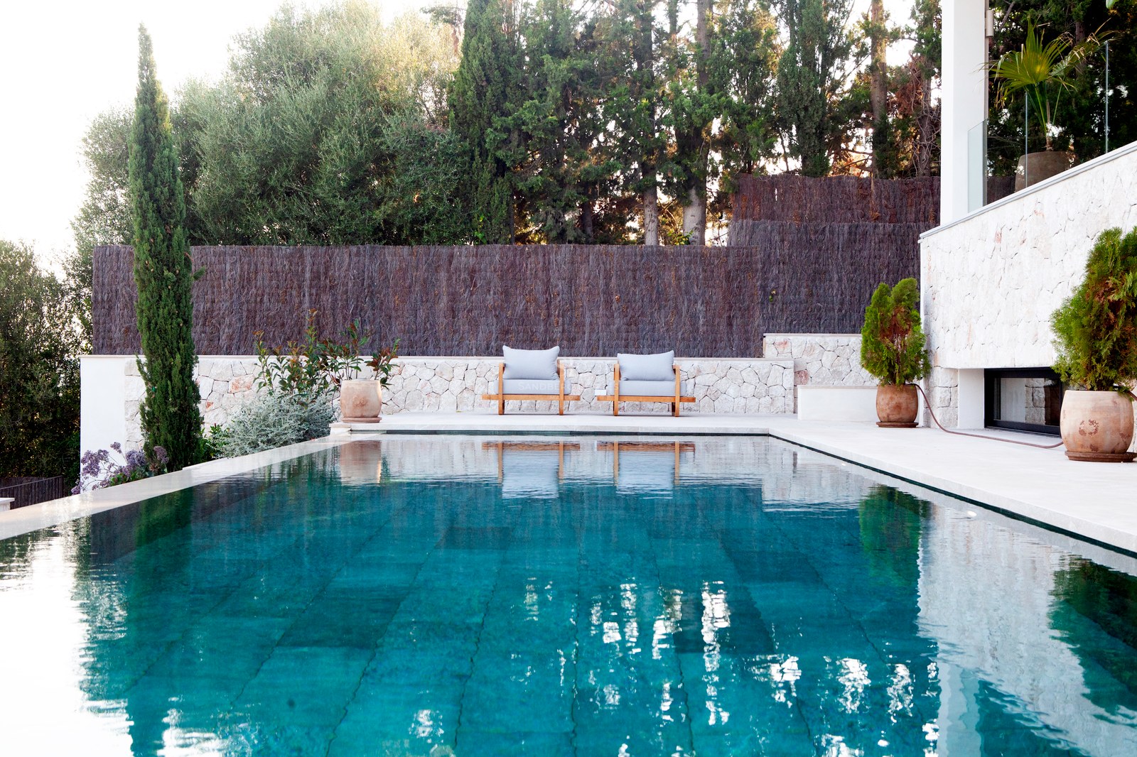 Türkisfarbener Infinity-Pool mit Natursteinumrandung, Holzliegen und üppigem mediterranem Garten.