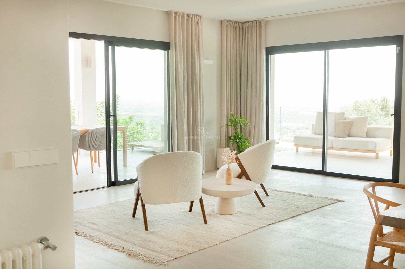 Modernes Wohnzimmer mit Bouclé-Sesseln, Steinboden und großen Glasschiebetüren, die auf eine sonnige Terrasse führen.
