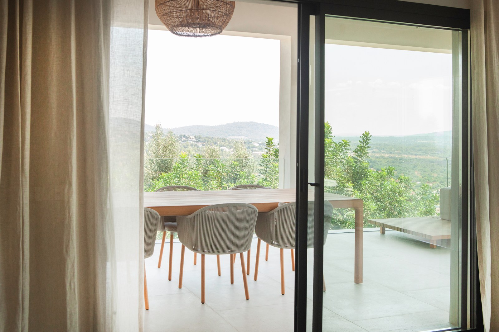 Blick durch Glastüren auf eine überdachte Terrasse mit Holztisch, Korbstühlen und Berglandschaft.