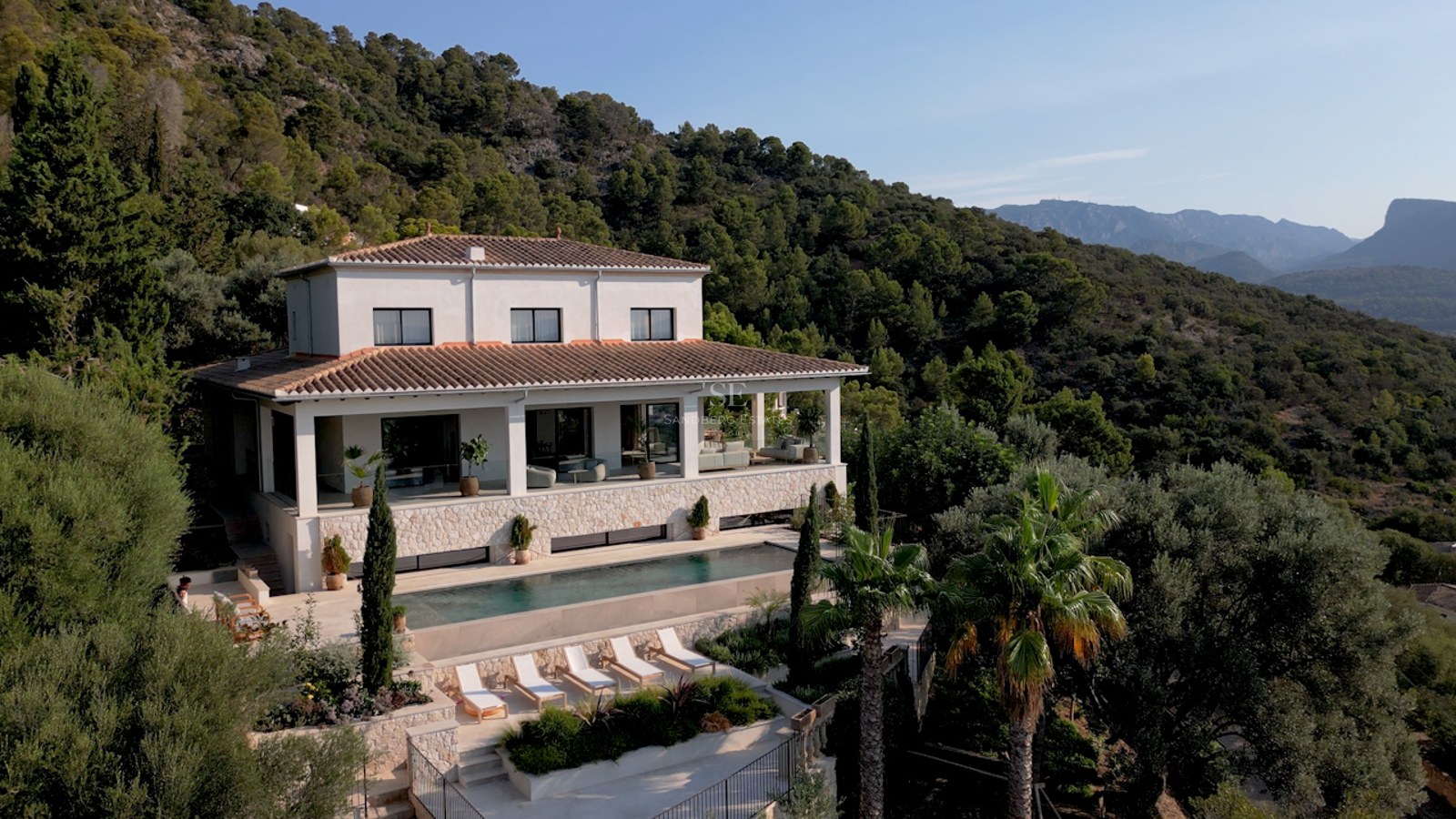 Erhöhte Ansicht einer weißen Villa mit Pool, Sonnenliegen und üppiger grüner Berglandschaft im Hintergrund.