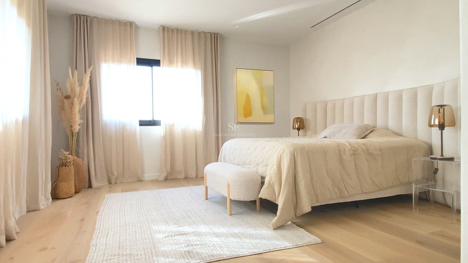 Helles Hauptschlafzimmer mit hellem Holzboden, raumhohen Vorhängen und gepolstertem Kopfteil in neutralen Tönen.