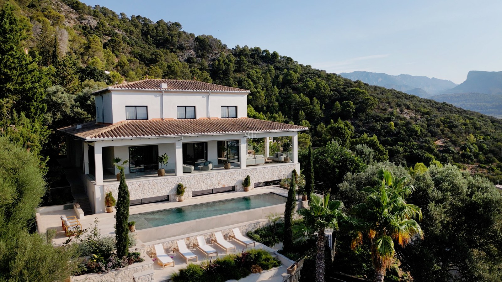 Luftaufnahme einer weißen zweistöckigen Luxusvilla mit Pool, Steinterrasse und üppiger Bergkulisse.