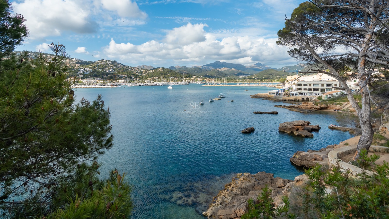 Vista elevada de una bahía mediterránea con yates, costa rocosa y montañas bajo un cielo azul con nubes.