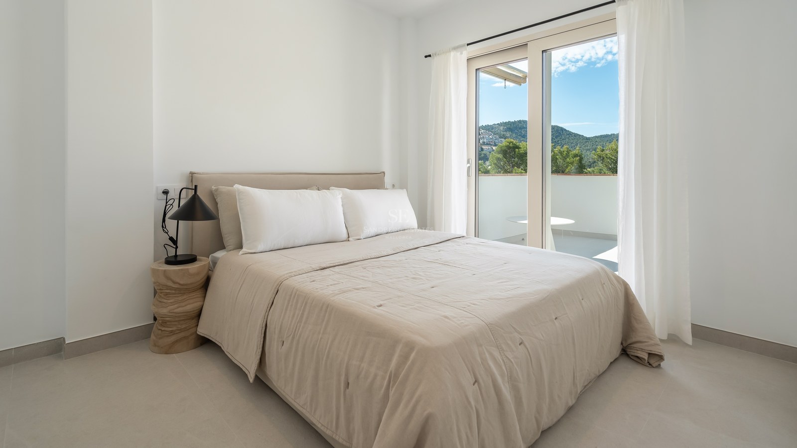 Dormitorio moderno con ropa de cama beige y puerta de cristal hacia un balcón privado con vistas a la montaña.
