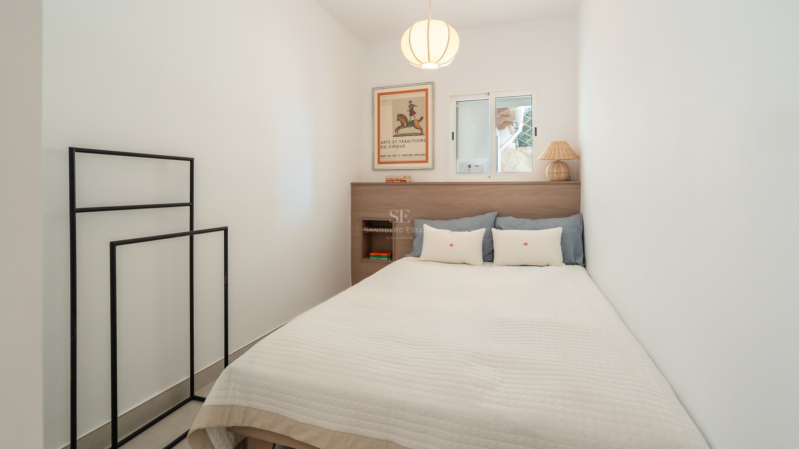 Dormitorio minimalista con cama blanca, cabecero de madera clara, ventana y lámpara de papel colgante.