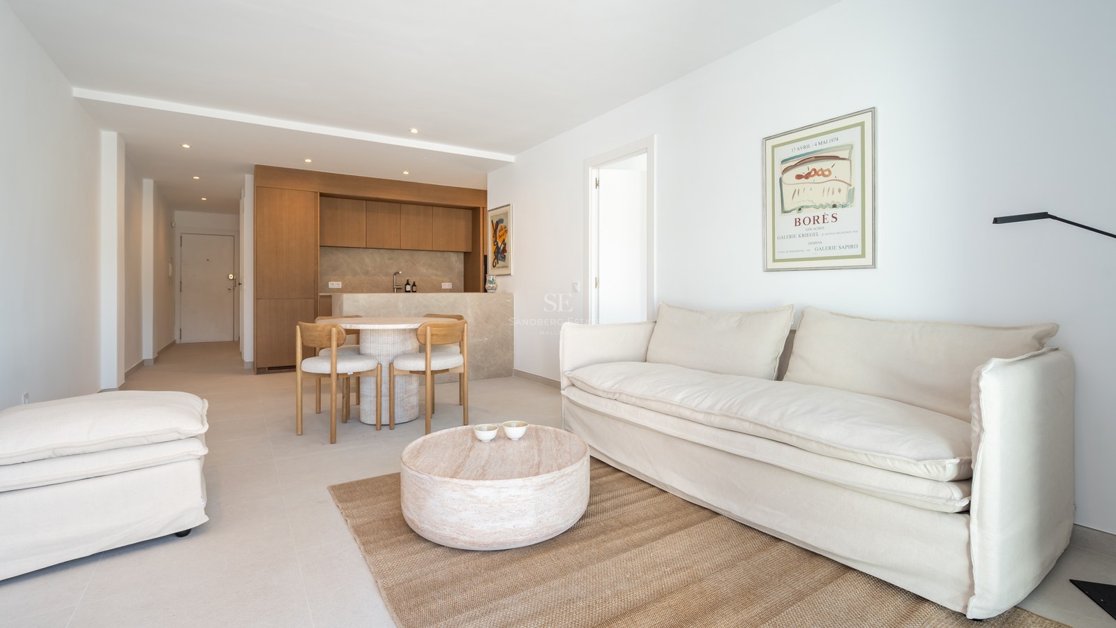 Salón luminoso con sofá de lino blanco, mesa de centro de madera redonda y cocina integrada al fondo.