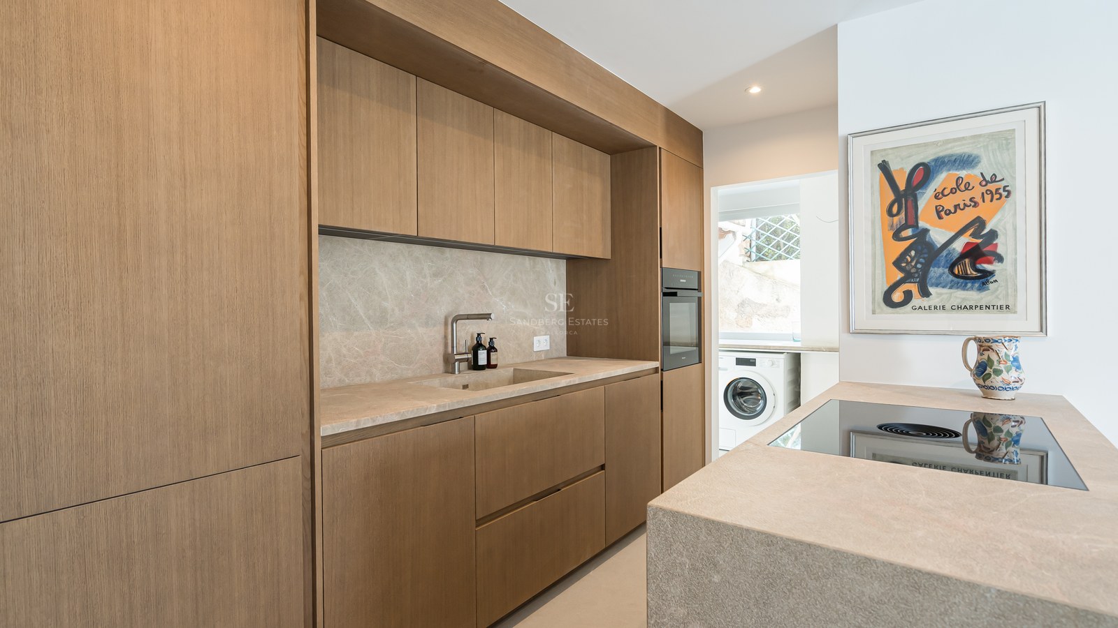 Cocina moderna con armarios de roble sin tiradores, encimera de piedra, horno integrado y zona de lavandería al fondo.