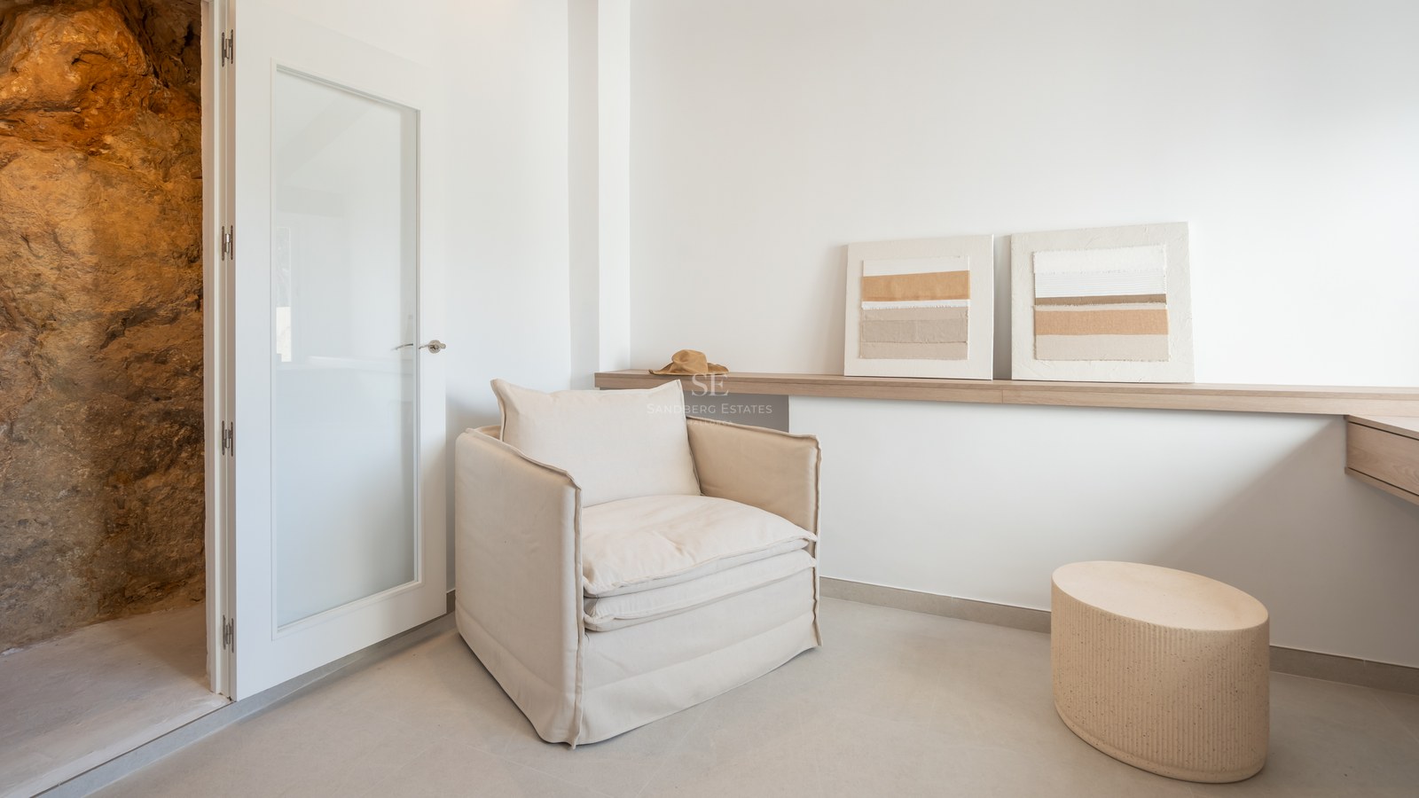 Sillón de lino beige junto a una estantería de madera clara con cuadros abstractos en una habitación luminosa.