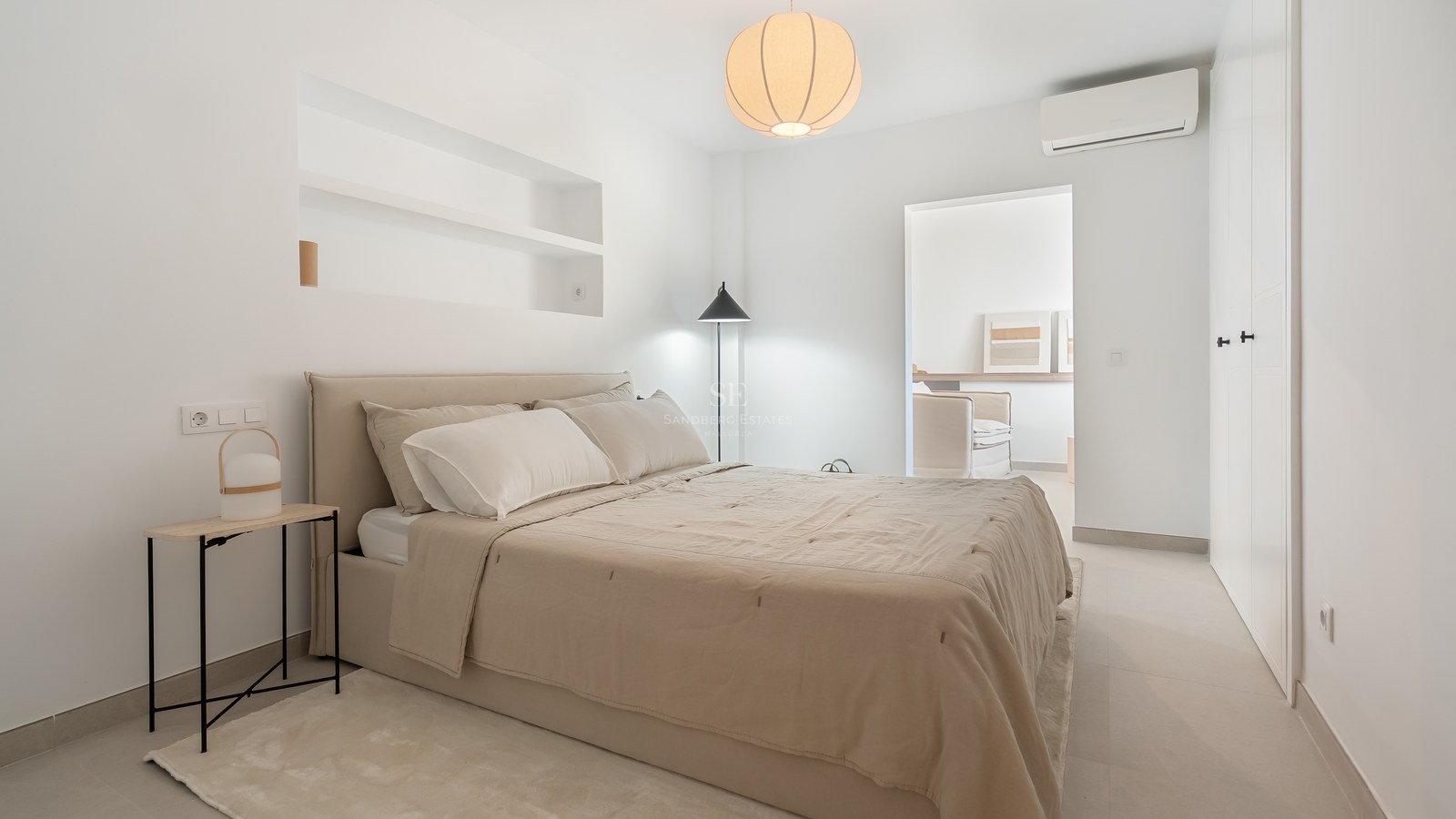 Dormitorio moderno con cama beige, estanterías integradas en la pared, lámpara de papel redonda y suelos claros.
