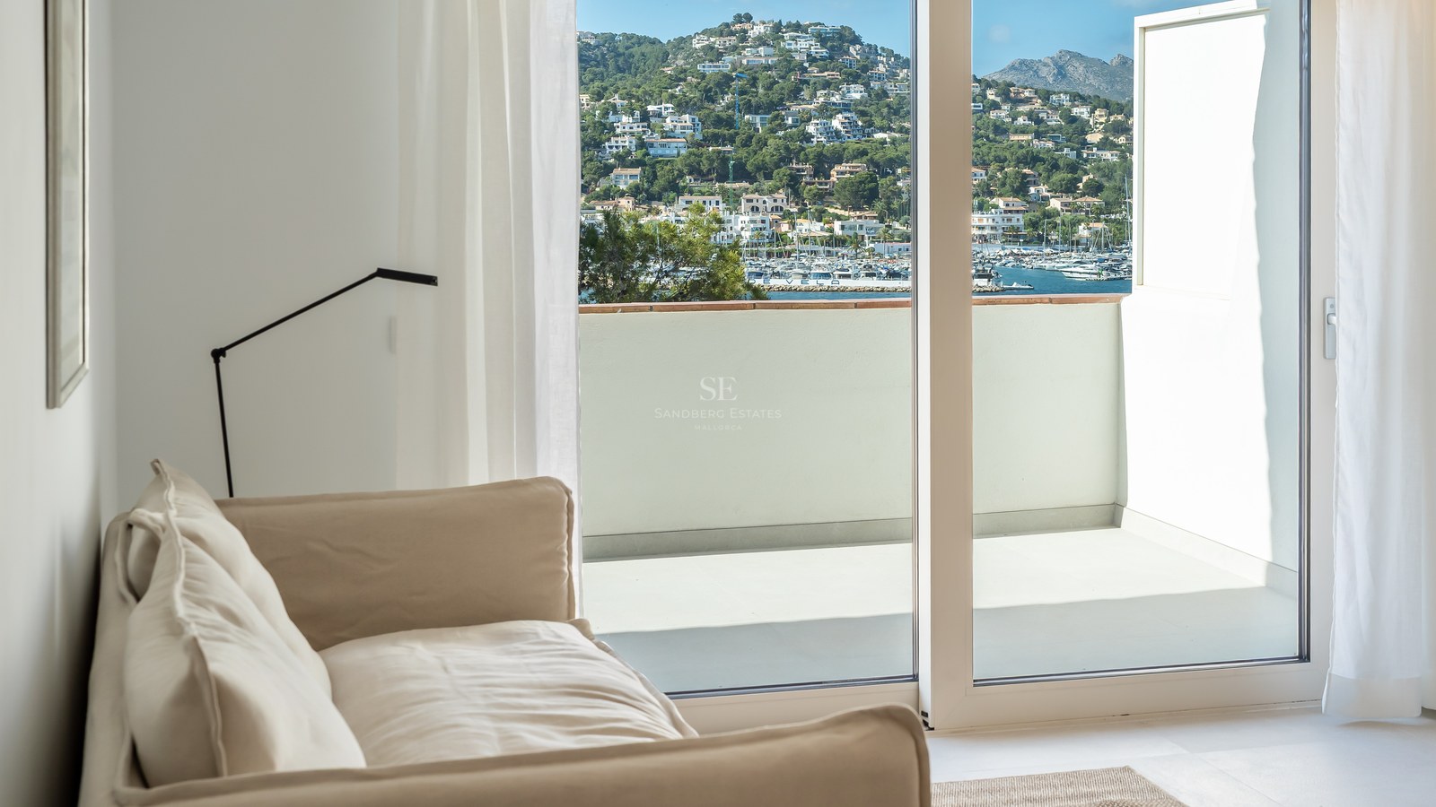Interior con sofá beige junto a ventanales de suelo a techo con vistas a un pueblo costero y puerto deportivo.