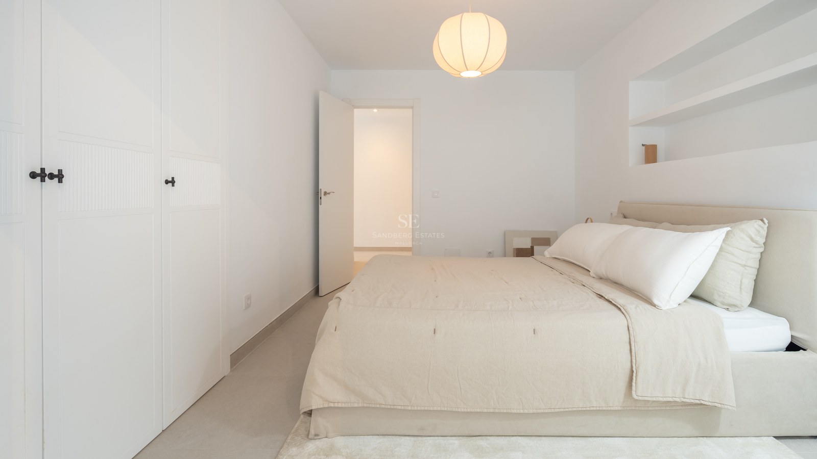 Un dormitorio luminoso y minimalista con una cama grande, sábanas blancas, una lámpara colgante redonda y una puerta abierta.