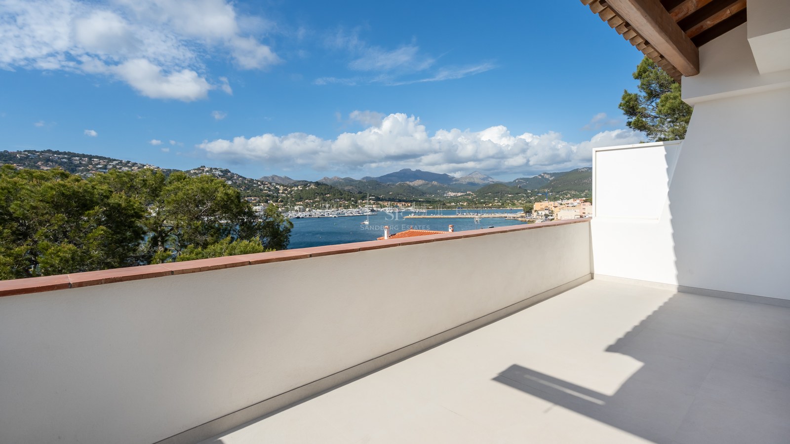 Amplia terraza blanca con vistas a un puerto deportivo con veleros, rodeada de colinas verdes y montañas.