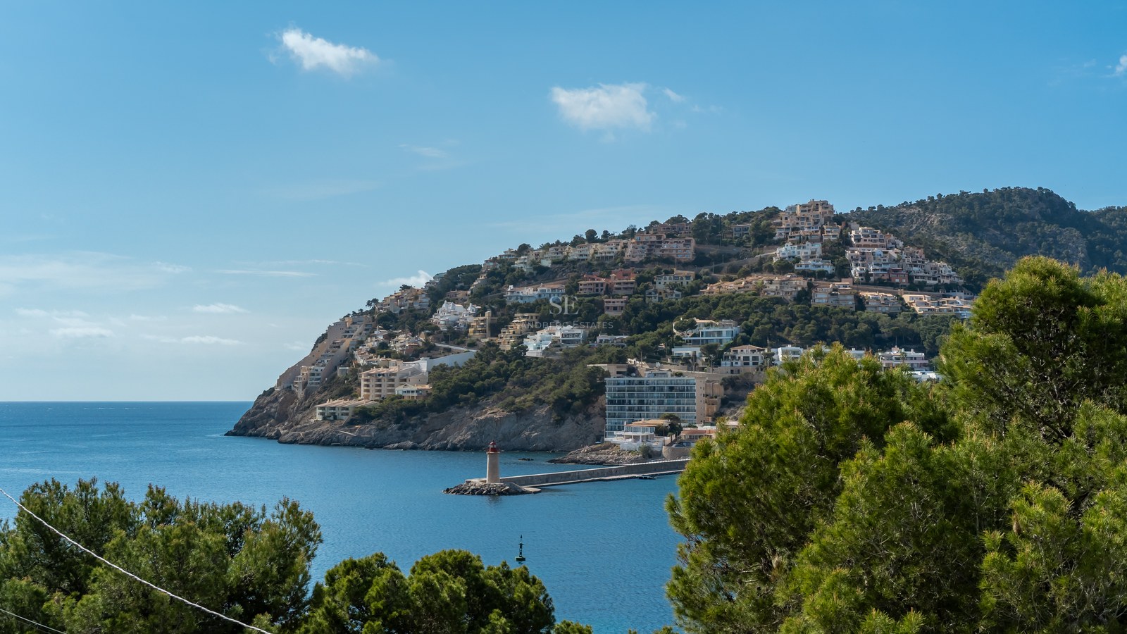 Paisaje mediterráneo con villas en la colina frente a una bahía azul con faro y pinos en primer plano.