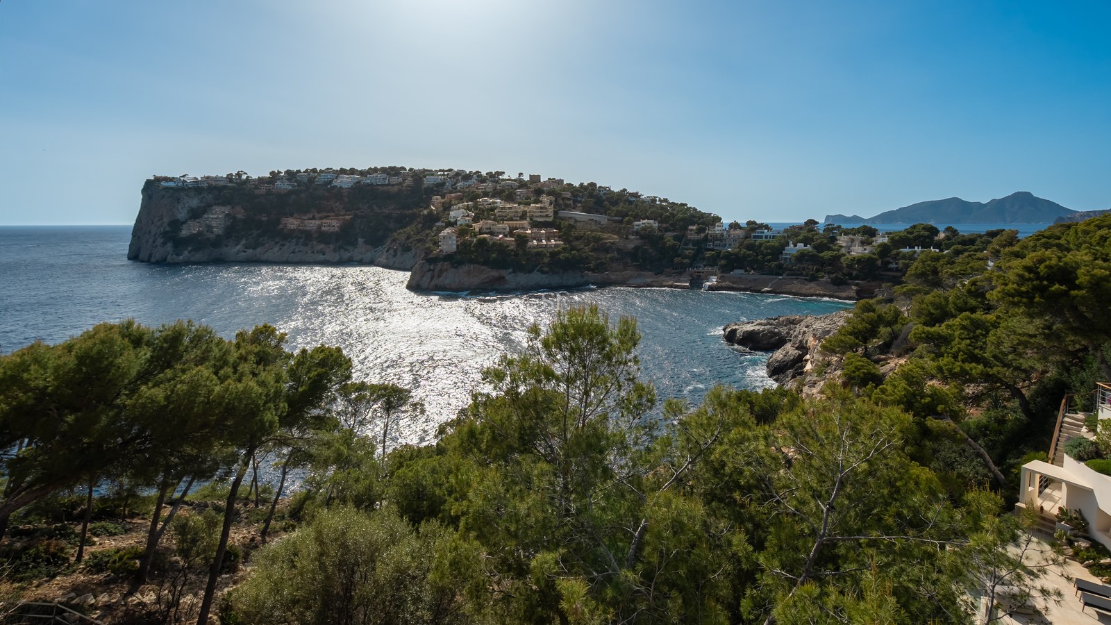 Baie côtière avec des eaux turquoise, des falaises rocheuses et des pins sous un ciel bleu clair.