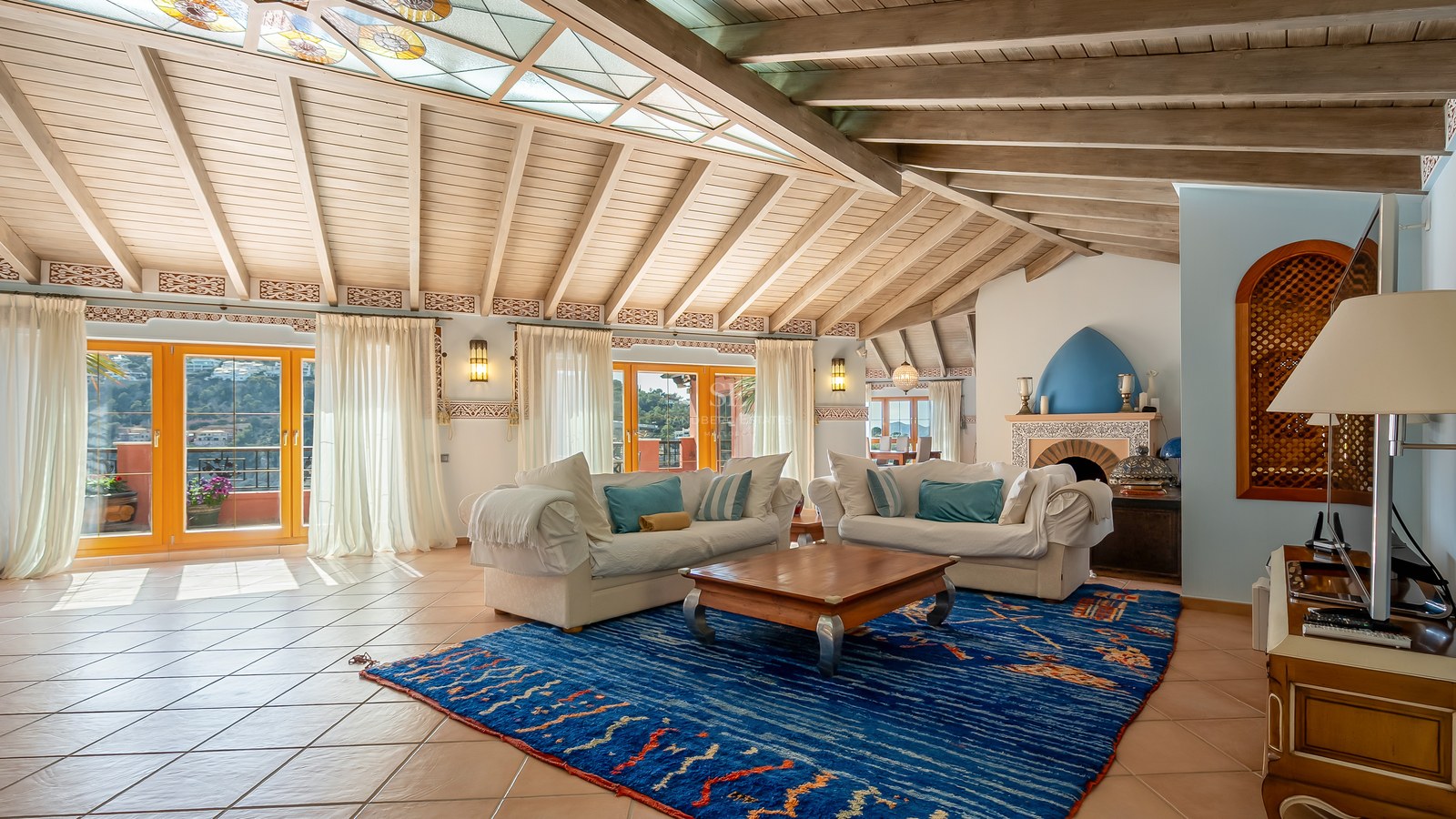Grand salon avec poutres en bois apparentes, sol en terre cuite, canapés blancs et un grand tapis bleu à motifs.